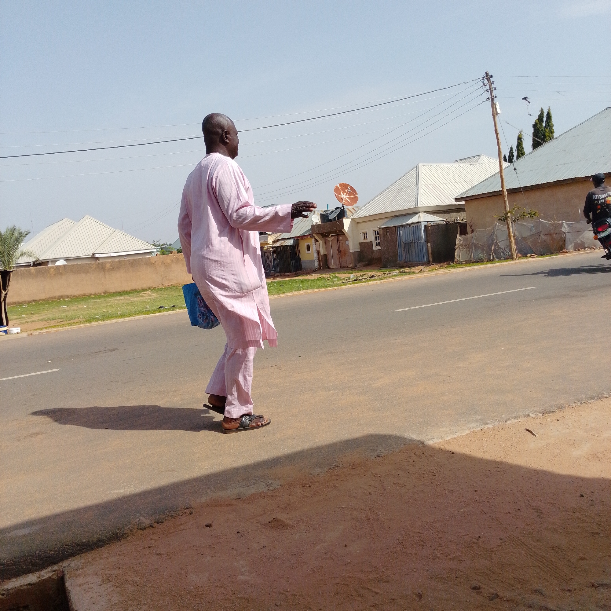 An adult male, facing away from the camera and towards the right, is walking on a paved road. He is wearing a light pink, striped traditional outfit and dark sandals. In his left hand, he carries a blue bag. He is walking past a line of houses with grey, sloped roofs and beige walls. A satellite dish is visible on top of one of the houses. Electrical wires crisscross the sky, supported by wooden poles. The road surface is asphalt, with a lighter-colored dirt shoulder. In the background, a motorcycle with a rider is also visible on the road. The scene appears to be in a residential area, likely within the city of Gombe, Nigeria, given the architectural style of the houses and the presence of satellite dishes, which are common in the region. The sky is clear and bright, indicating daytime.
