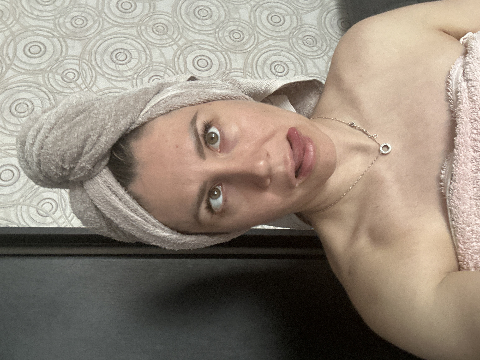A young woman with a towel wrapped around her head and shoulders is captured in a selfie. She is lying down, with her head tilted and her tongue sticking out, looking at the camera with a playful expression. Her makeup is visible, with eyeliner and mascara accentuating her eyes. A delicate silver necklace with a circular pendant adorns her neck. The background features textured wallpaper with a circular pattern and a dark surface, possibly a piece of furniture. The lighting suggests it could be daytime indoors. The woman is wearing a pink towel over her shoulder, complementing the light-colored towel on her head. The overall impression is a lighthearted and casual moment.