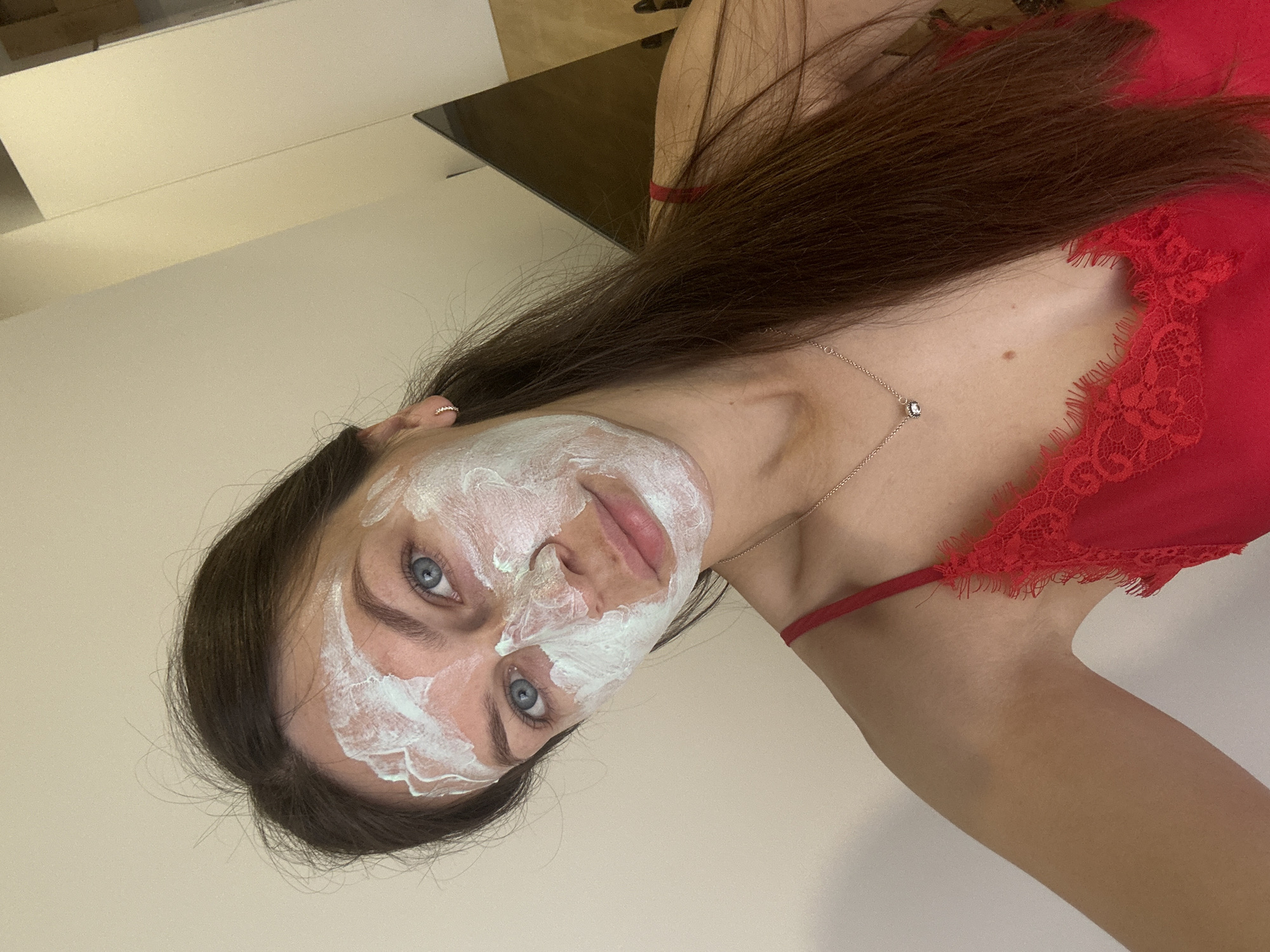 A young woman with a white facial mask is angled in the frame. She has long brown hair and blue eyes. She is wearing a red lace top. A silver necklace is visible. The background appears to be a plain, light-colored wall. Based on the request, the location is Donji Pobori, Montenegro, but there are no visual cues in the image to confirm this.