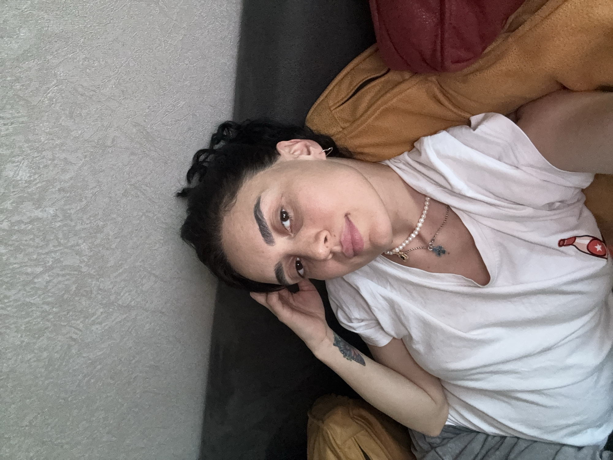 A young woman with dark curly hair and light skin is lying on a sofa, looking directly at the camera. She is wearing a white t-shirt with a small red and white embroidered bottle on the left side of the chest. She has a pearl necklace with a cross pendant and a silver hoop earring in her left ear. There is a tattoo on her left forearm. The background appears to be a textured wall and a dark gray sofa. Cushions in mustard yellow and red are visible to her right and left, suggesting she is indoors. The scene does not provide any specific indicators of location within Tbilisi, Georgia.
