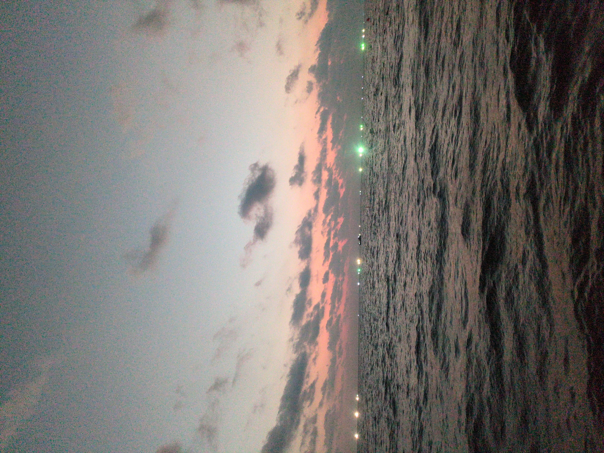 The image depicts a view of the sea at sunset from Phú Quốc, Vietnam. The sky is a mix of gray and pink hues with scattered clouds. The water surface is dark and rippled. In the distance, a line of lights, possibly from boats or the coastline, is visible. There are no people present in the image.