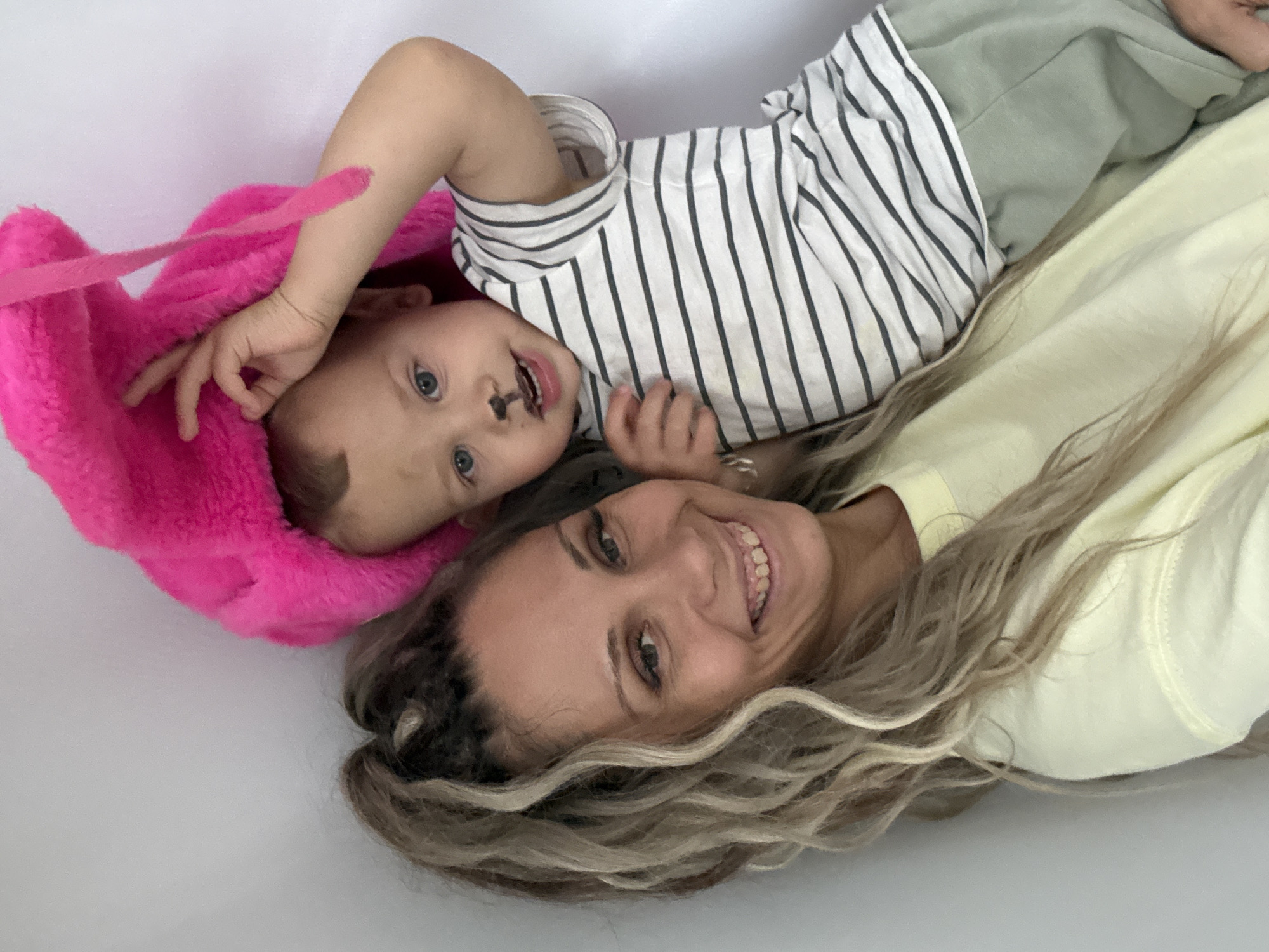 A close-up, high-angle selfie shows a woman and a young child against a plain, light-grey background. The woman is on the left, holding the child. She has long, blonde, curly hair pulled into a high ponytail and is smiling at the camera. She is wearing a light yellow sweatshirt. The child is on the right, wearing a striped t-shirt and light green pants. The child has a black smudge on his nose and above his upper lip, resembling a mustache or whiskers. He is wearing a large, bright pink fuzzy hat that partially obscures his head. The child is looking slightly off-camera and has his right hand raised, touching the hat. The lighting is bright and even, suggesting an indoor setting.