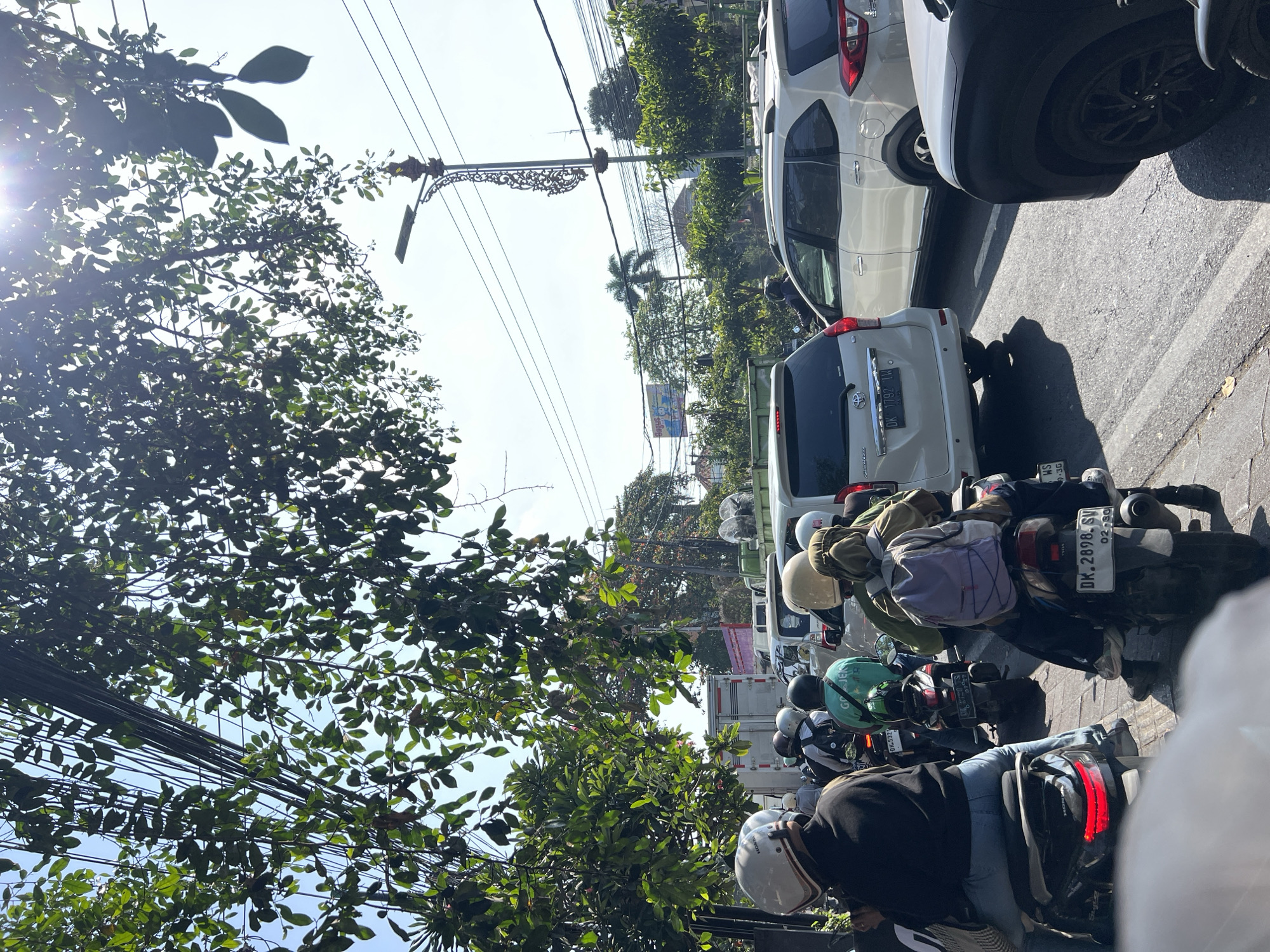 The image depicts a traffic jam on a sunny day in Denpasar, Indonesia. The foreground is dominated by the rear of several motorcycles, with riders wearing helmets. One motorcycle in the center has a blue backpack on the passenger seat. A white SUV is visible in the middle ground, with a truck behind it. The background shows more traffic and lush green trees. Overhead, power lines crisscross the sky, and a street lamp with decorative detailing stands tall. The scene suggests a typical urban street environment in Indonesia.