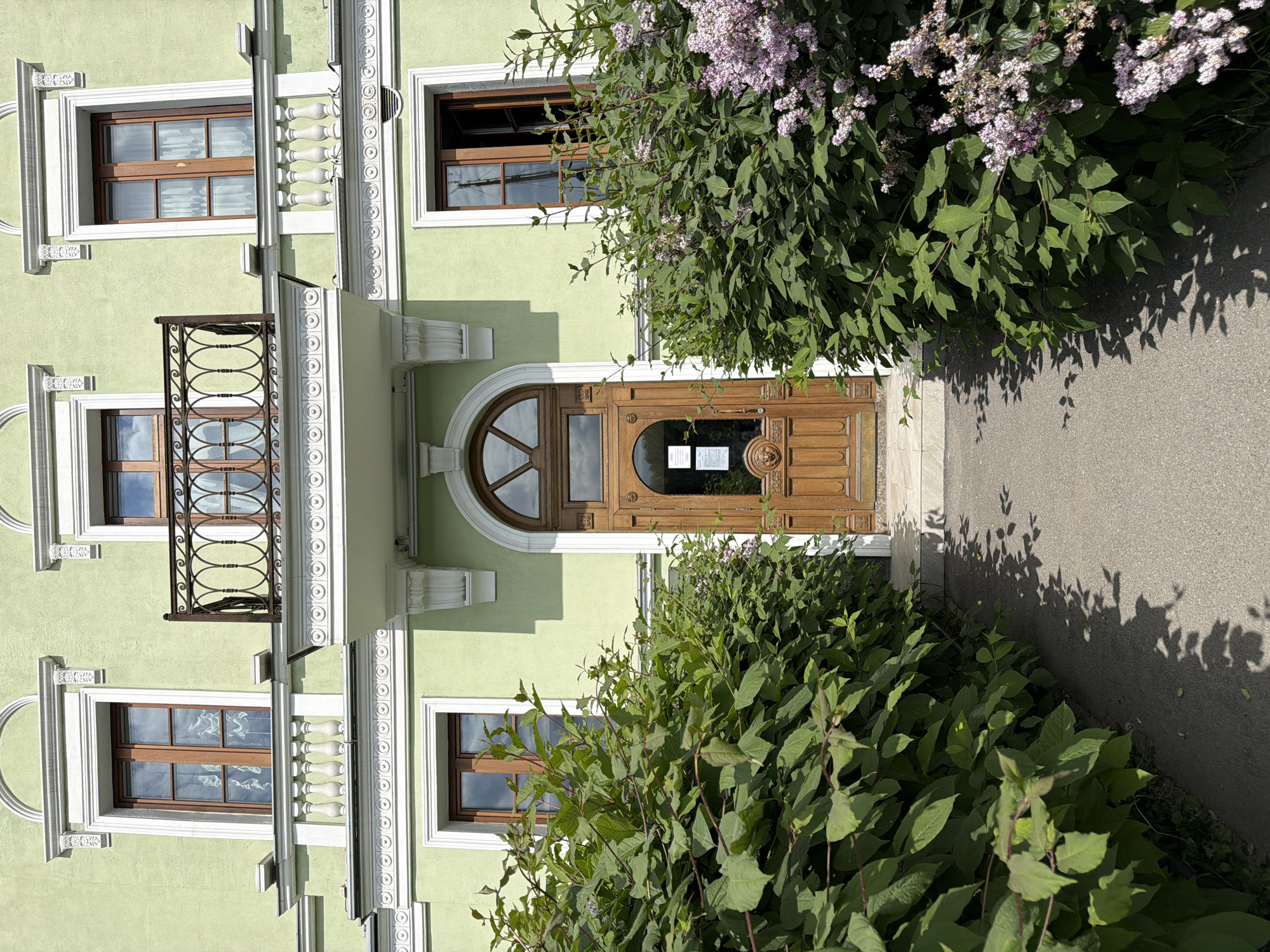 This image captures the facade of a light green building, likely located in Saint Petersburg, Russia. The building features ornate white architectural detailing around its windows and a prominent wooden main entrance door with an arched transom window above. Wooden window frames are visible in the multiple rectangular windows, some of which are partially open. Above one of the windows, there is a small, decorative balcony with an intricate black metal railing.
Lush green bushes, some adorned with clusters of light purple flowers, line the paved pathway leading to the entrance, partially obscuring the lower sections of the building. Strong, clear sunlight casts distinct shadows of the foliage onto both the pathway and the building's facade, indicating a bright, sunny day. A small, white rectangular sign is affixed to the main door, but the text on it is illegible. The scene is static, with no people or activities visible.