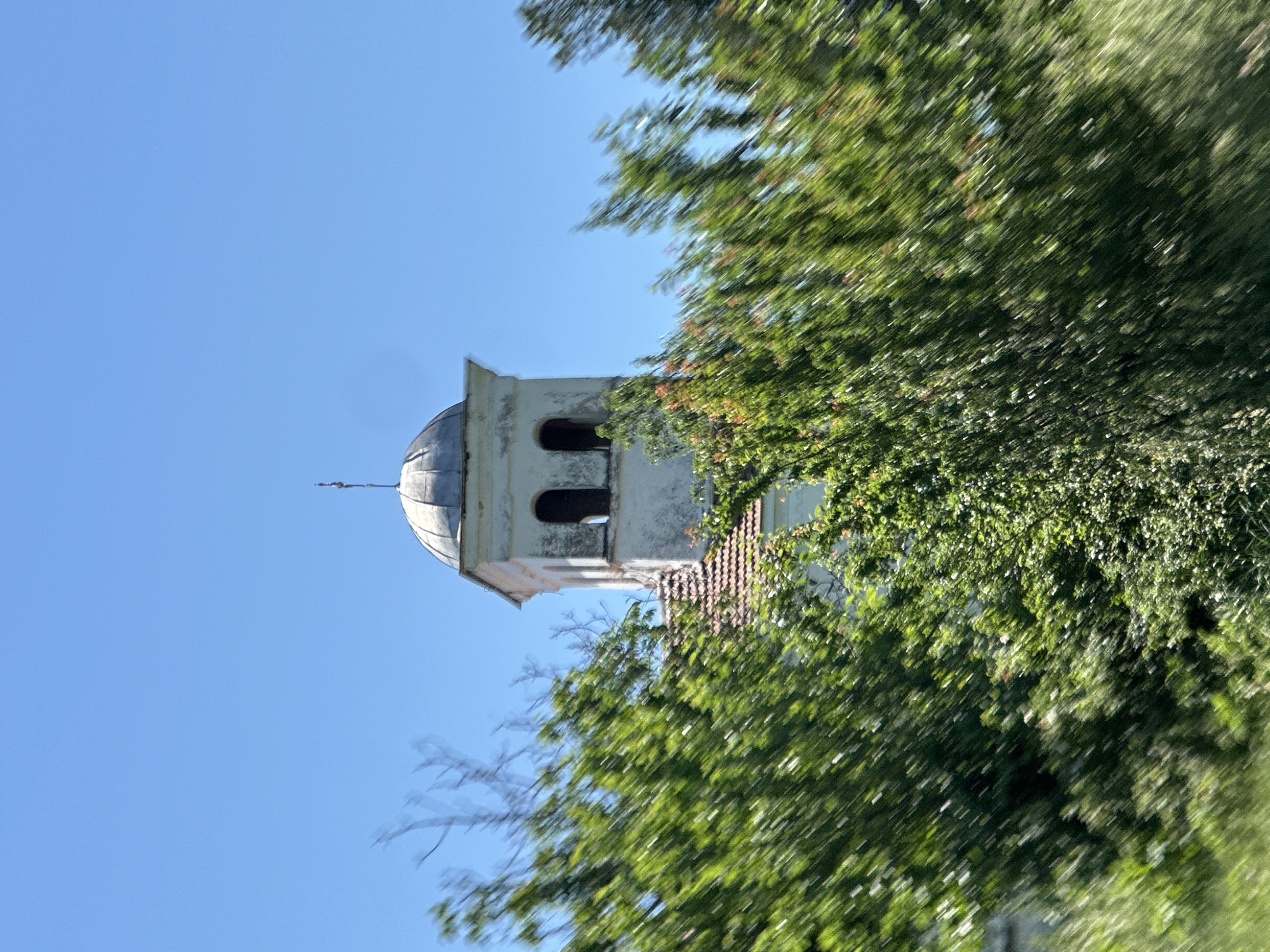 An upward shot showing a tower with a dome-shaped top of a church, partly hidden by green foliage, against a clear blue sky. The church is located in Slavyani, Bulgaria. The tower is off-white and appears to have two arched windows. A thin cross is seen at the very top of the dome. The foliage is blurred, suggesting movement.