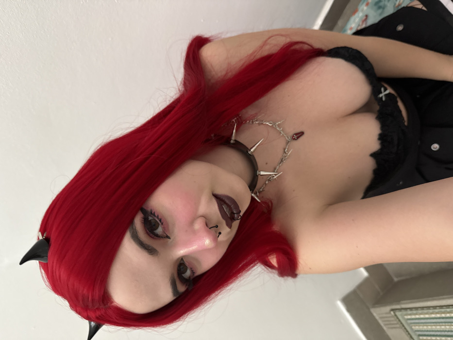 A woman with bright red hair and fair skin tilts her head towards the camera. She has dark eye makeup and dark lipstick, a nose ring, and is wearing a studded choker. Her outfit appears to be gothic-inspired, with a black lace top visible. Small devil horns are placed on her head. The background is a plain white wall.