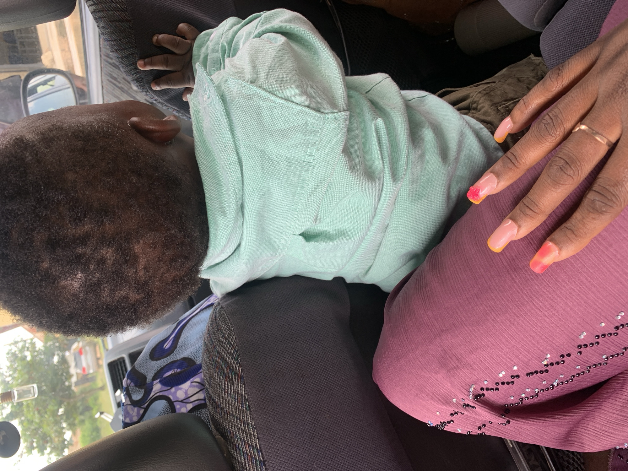 The image shows a close-up view of a child and a woman's hand inside a vehicle. The child, facing away from the camera, is wearing a light mint green collared shirt and brown shorts. The child's head is turned to the left, with their ear visible. The woman's hand, with long, bright pink and orange gradient nails and a ring on one finger, rests on the woman's lap, which is covered by a dark pink, pleated fabric adorned with black and silver beads. In the background, through the car window, trees and another vehicle are visible. The scene appears to be from a street in an urban or suburban area, though specific details of the location within the city of Unknown, Unknown are not discernible. The overall lighting suggests daytime.