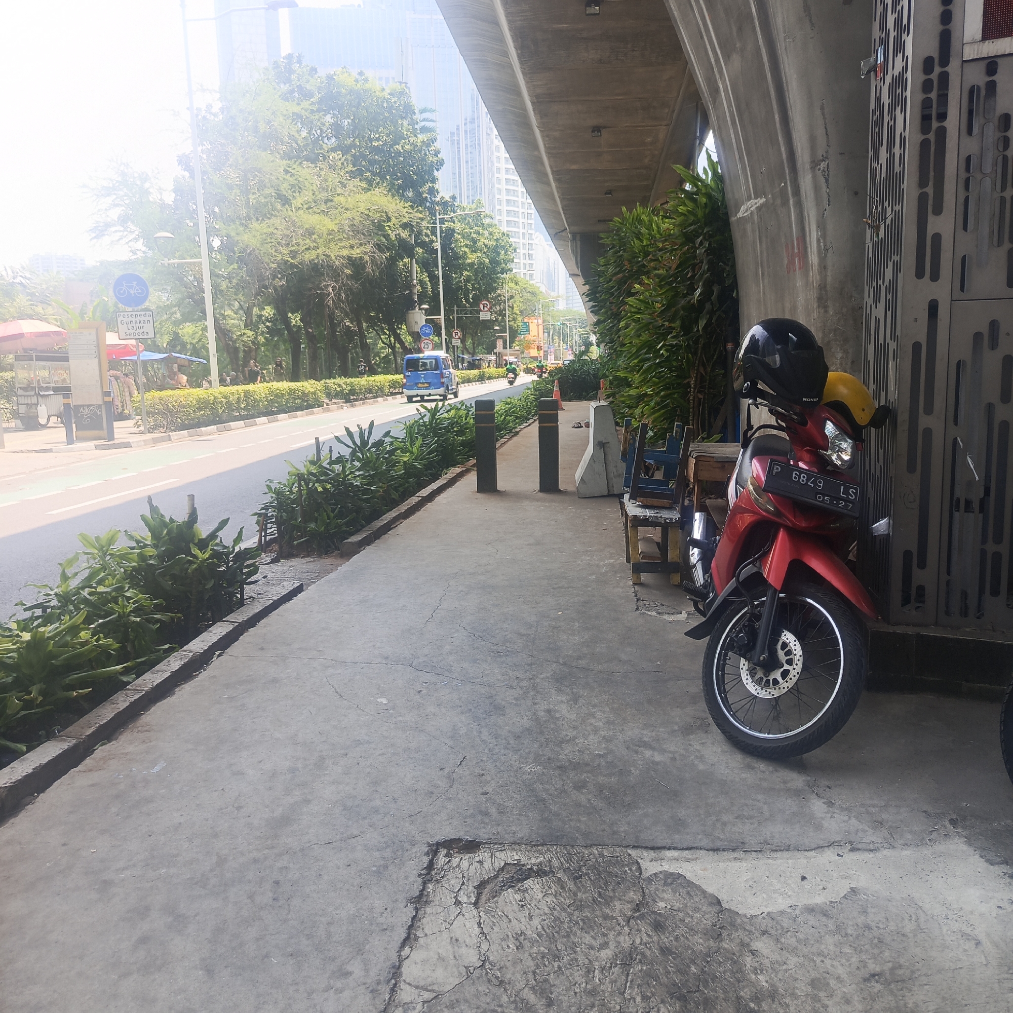 The image captures a street scene in South Jakarta, Indonesia, under an elevated structure, likely an overpass or flyover. A red motorcycle with a black helmet and yellow visor is parked on the right side of the frame, against a textured metal partition. The motorcycle's license plate reads "P 6849 LS" with "05-27" below it. A sidewalk, made of concrete with visible cracks and repairs, runs along the left side of the frame. On the sidewalk, two dark cylindrical bollards are placed side-by-side. Beyond the sidewalk, a road is visible with a dedicated bicycle lane marked by white lines. A blue minivan and several other vehicles, including motorcycles, are driving on the road. Lush green foliage lines the side of the road and the sidewalk. In the background, tall buildings typical of a city skyline are present. The overall lighting suggests it is daytime, with bright sunlight creating some lens flare in the upper left corner. Several wooden chairs are stacked on the right side of the sidewalk near the motorcycle. A sign with a bicycle symbol and text in Indonesian, "Pesepeda Gunakan Lajur Sepeda" (Cyclists Use Bicycle Lane), is visible on the left side of the road.