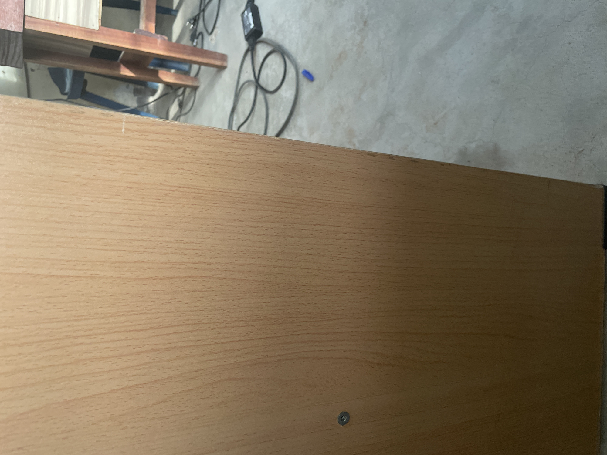 The image shows a close-up of a light-colored wooden surface, possibly a cabinet or desk, with a visible screw head on the left side. To the right of the wooden surface, a concrete floor is visible with several electrical cords, a black power adapter, and a blue pen. In the background, parts of wooden furniture, possibly a table and chairs, can be seen. The overall environment appears to be indoors, in what could be a workshop or a storage area. The image does not provide any information about the city of Jalingo, Nigeria.