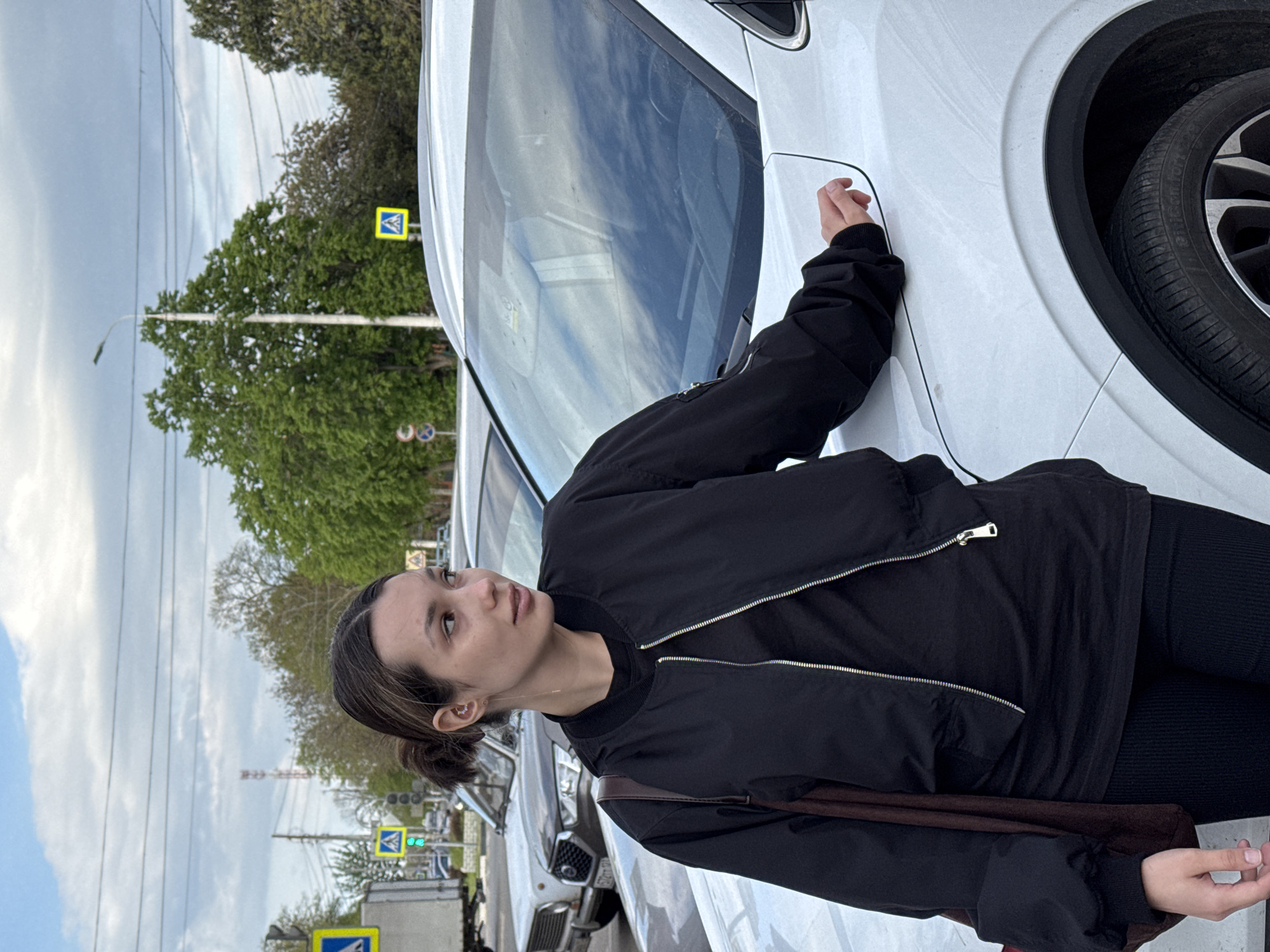 The image is vertically oriented, featuring a young woman standing beside a white car on what appears to be a street or parking area during daytime under an overcast sky.
The main subject is a young woman, seen from the waist up, facing mostly forward but looking slightly upwards and to her right. She has dark hair styled in a bun or ponytail. She is wearing a black bomber jacket with silver zippers, a black t-shirt, and black leggings or trousers. A brown strap, possibly from a bag, is visible over her right shoulder. Her left hand rests gently on the white body of the car. Her expression is neutral and contemplative.
She is standing next to a white vehicle, likely a car or SUV, with part of its front wheel and tire visible on the right side of the frame. The car's windshield reflects the sky and some background elements.
The background consists of several elements:
- Lush green trees line the street, suggesting a temperate climate.
- A light grey, overcast sky fills the upper portion of the frame.
- Power lines stretch across the sky.
- A street lamp post is visible amidst the trees.
- Multiple yellow diamond-shaped pedestrian crossing signs are discernible in the middle ground, indicating a street or urban setting.
- Other cars are parked or driving in the distance, one of which shows a partially visible license plate with the numbers "572".
The overall scene depicts an ordinary moment in an outdoor urban or suburban environment.