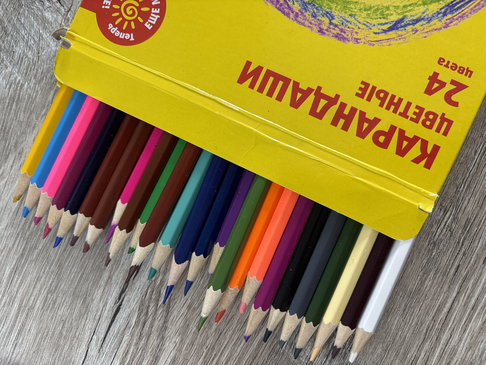 The image shows a collection of colored pencils fanned out on a wooden surface. The pencils are mostly within a yellow box that has Russian text on it, which appears to read "КАРАНДАШИ ЦВЕТНЫЕ 24 цвета", translating to "COLORED PENCILS 24 colors". The wooden surface has a prominent grain pattern. There are no people or animals in the image. The scene is static and indoors. The location of this image is not specified and cannot be determined from the provided visual information, therefore it cannot be located in Sochi, Russia.