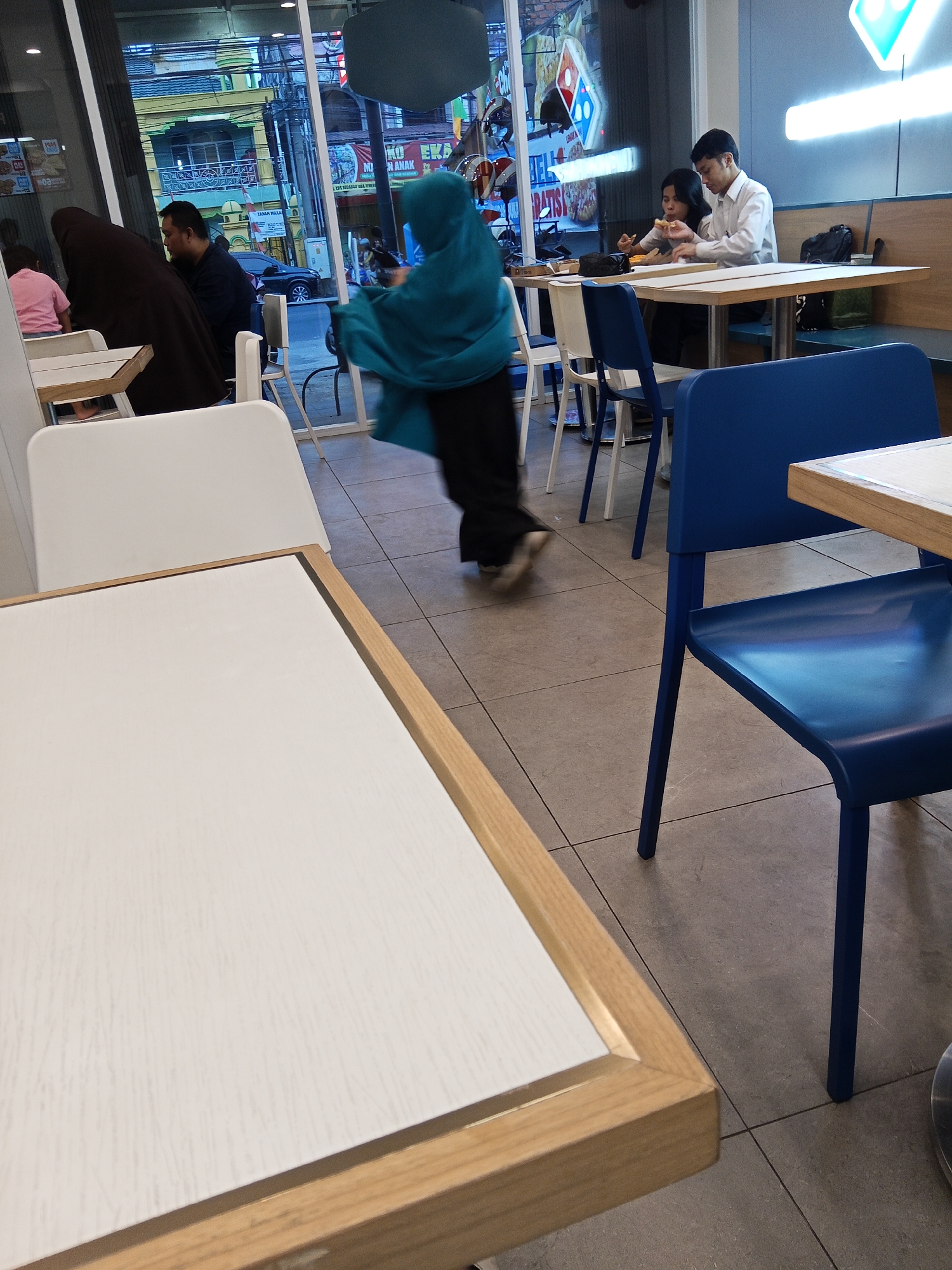 The image depicts the interior of a Domino's Pizza restaurant, possibly located in Lubuklinggau, Indonesia, based on contextual information. In the foreground, a white tabletop with a light wood trim is visible. Behind it, a blue plastic chair with dark legs is positioned. Further in the background, through a large window, a street scene is visible with buildings and a black car.
Inside the restaurant, several patrons are present. Two individuals, a man in a white shirt and a woman, are seated at a table, eating. A person wearing a teal hijab and dark clothing is walking past the tables. Another patron, also wearing a dark hijab, is seated at a table with another person. White chairs and tables are also present. The restaurant features a prominent Domino's Pizza sign with neon lighting. The flooring is tiled in a gray, grid pattern.