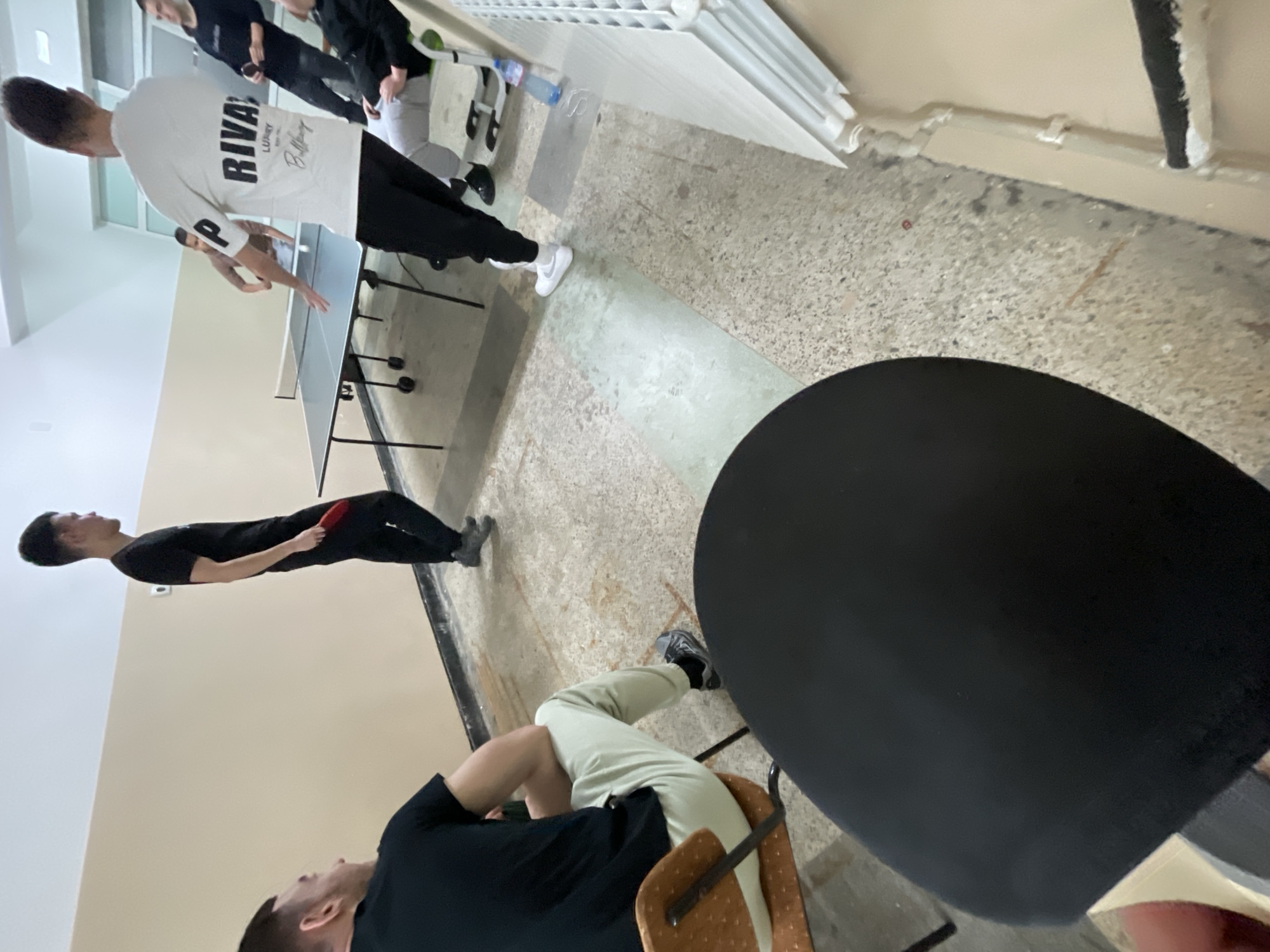 A group of people are playing ping pong indoors. One person is hitting the ball while another observes. A seated individual is in the foreground. The location is possibly in Zlatograd, Bulgaria, but there is no supporting evidence for this.
