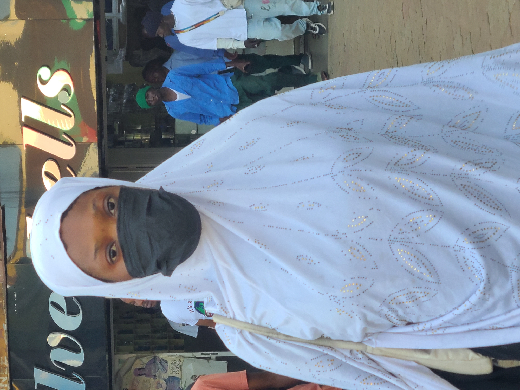 The image captures a street scene in front of a business with a prominent sign reading "Silverlls" in Jos, Nigeria. A woman wearing a white hijab and a black face mask is the central figure, positioned in the foreground and looking directly at the camera. Her white hijab is adorned with a pattern of small golden leaf-like designs. A light-colored bag hangs from her left shoulder. Behind her, the entrance of the business is visible, with shelves stocked with items. Several individuals are seen in the background, including a man in a blue shirt and green pants, and another in a white t-shirt and light blue jeans. The environment appears to be an urban commercial area, with a paved ground surface and other shops or buildings visible in the distance. The lighting suggests it is daytime.