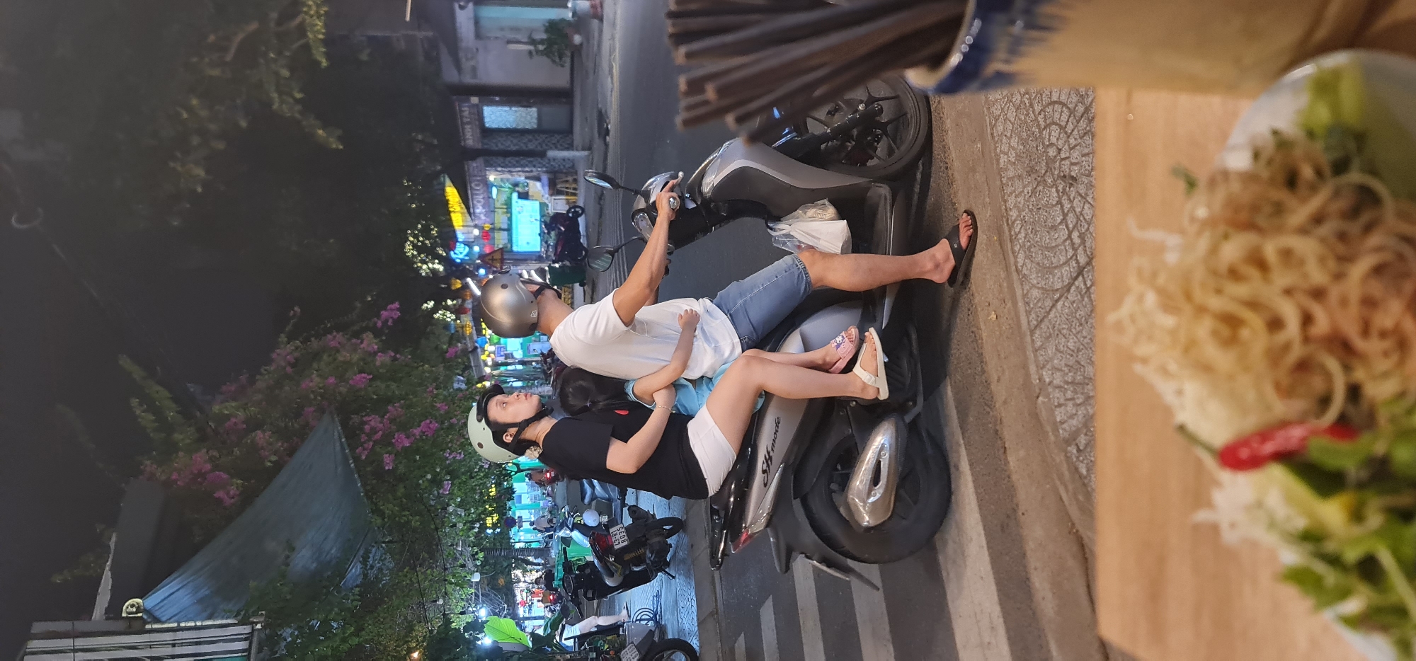 A medium-shot, medium-angle photograph captured at night depicts a family of three seated on a parked scooter in what appears to be Đà Nẵng, Vietnam. A man wearing a gray helmet and a white t-shirt is in the driver’s seat, his hand positioned on the scooter's handlebar. He is flanked by a woman in a black t-shirt and light green helmet. Sandwiched between them is a small child, dressed in a blue shirt. The man wears jeans and black flip-flops while the woman is dressed in white shorts and white sandals.
The scooter, an "SH Mode," is parked on the side of a street paved with gray stones. Additional scooters and pedestrians are visible in the background. Further back, one can see buildings with illuminated signage. A flower bed with pink flowers lines the edge of the sidewalk. Chopsticks in a ceramic container and a plate of noodles are visible in the extreme foreground, suggesting that the photo was taken at a restaurant or food stall.