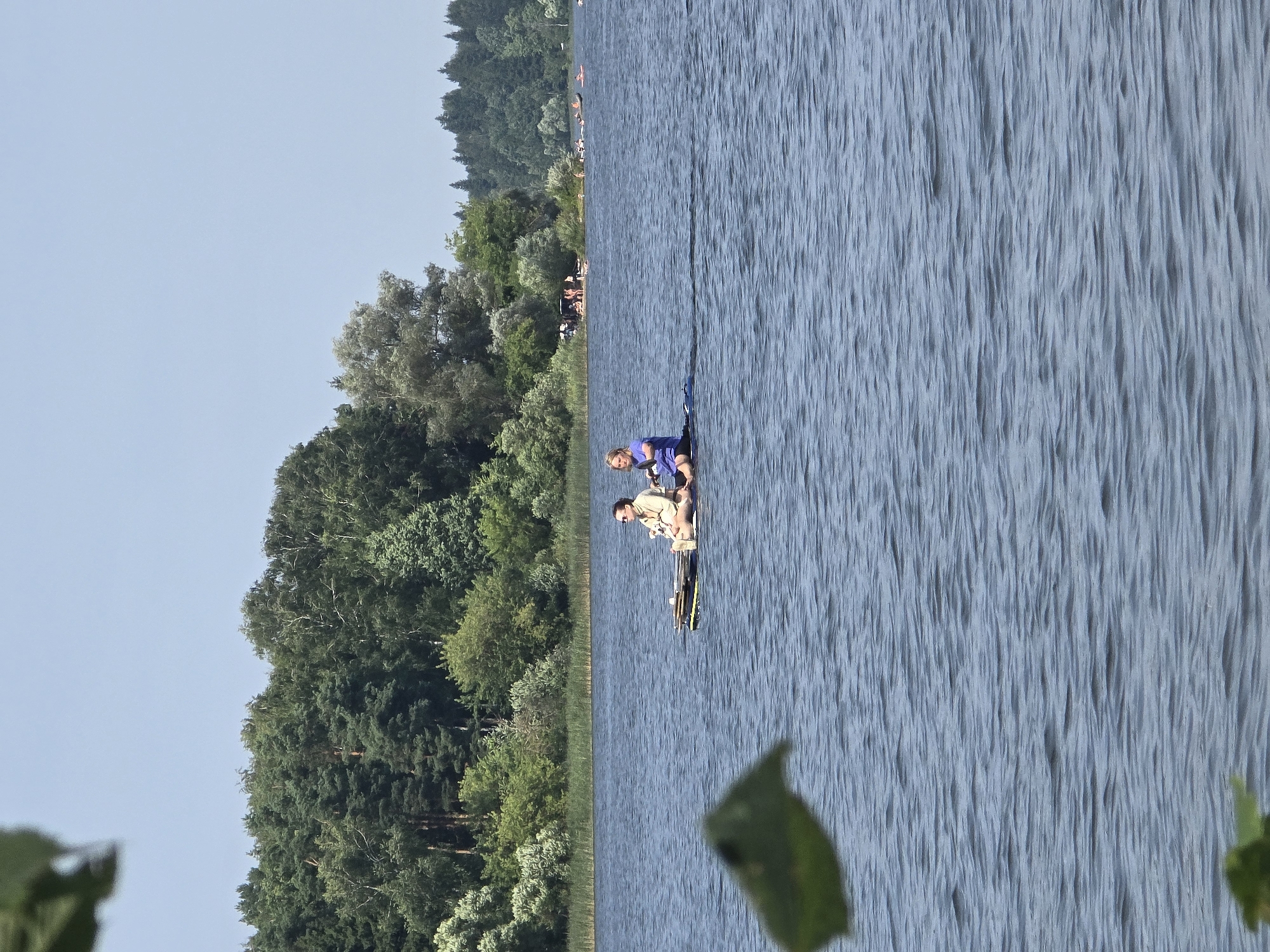 The image depicts a serene, wide-angle view of a calm body of water, likely a lake or reservoir, under a clear sky. In the foreground, out of focus, a large green leaf hangs from the top left corner, and another from the bottom left corner, suggesting a natural outdoor setting.
Dominating the mid-ground are two individuals on a raft or flat-bottomed boat. One person, wearing a light-colored jacket and dark pants, sits with their legs extended, holding a drink. Beside them, another person, dressed in a blue top and dark shorts, is actively paddling with a single oar, suggesting they are either propelling the raft or enjoying a leisurely paddle. The raft itself appears to be constructed from wood or a similar material.
In the background, a lush, dense forest lines the far bank of the water. The trees are a mix of various shades of green, with some lighter foliage interspersed. Further in the distance, along the shoreline, there are visible signs of human activity, including a cluster of people gathered on a beach or near the water's edge. More people can be seen scattered across the water on various flotation devices, some appearing to be swimming or engaged in water activities. The overall impression is one of a pleasant, recreational day in a natural environment. The location is identified as the settlement of Kuybyshev Hydroelectric Station, Russia, known for its water bodies and recreational opportunities.