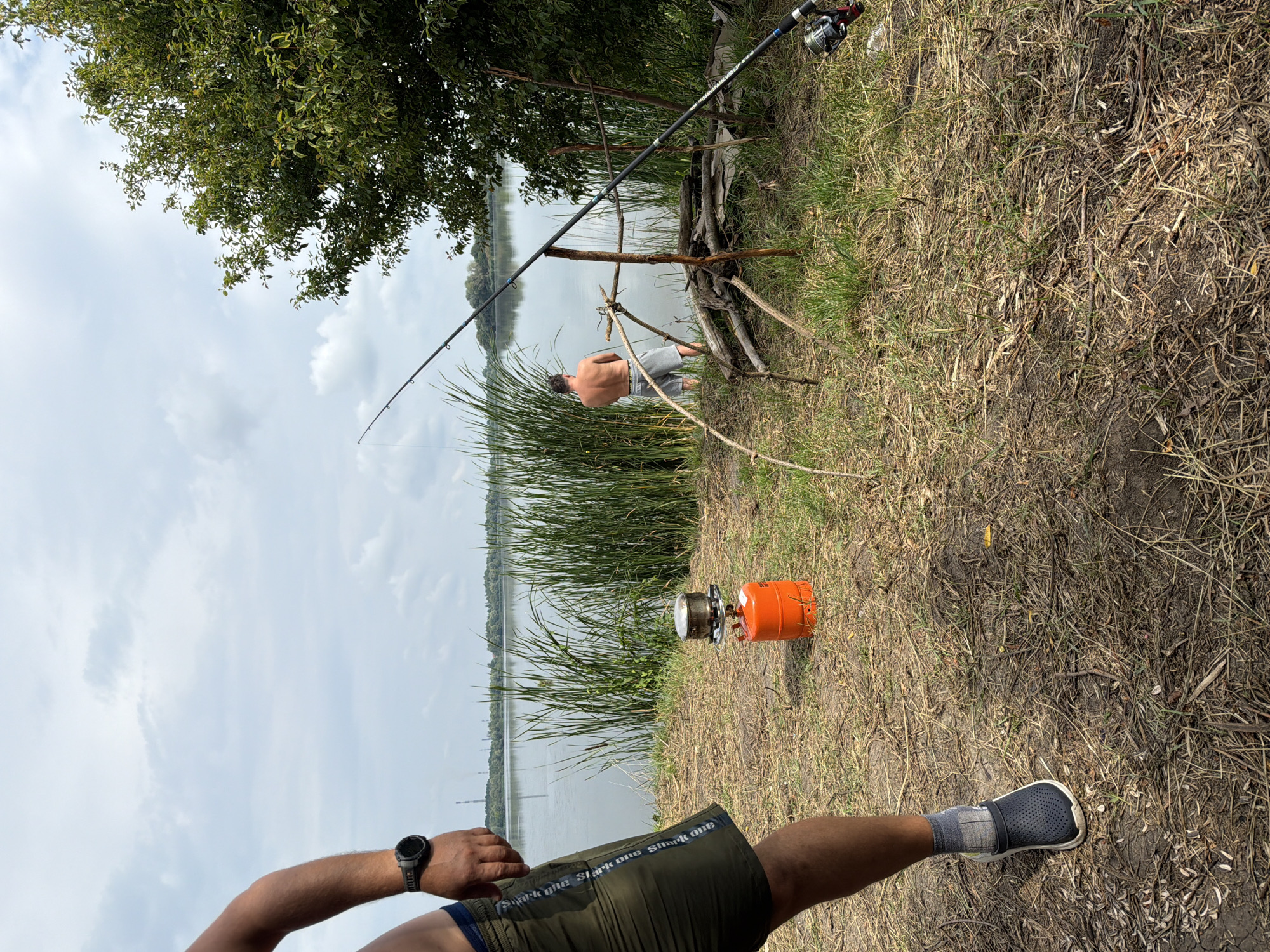 In Ruda, Ukraine, on a cloudy day, two men are by a body of water. The foreground shows the legs of one man wearing a grey sock and a dark grey clog, with green shorts and a watch on his left wrist. To his right, an orange portable stove with a pot on top sits on dry grass. The middle ground features a second man, shirtless and wearing grey shorts, standing near the water's edge facing away from the camera. Between him and the camera is a makeshift structure made of branches, with a fishing rod propped against it. The background displays a calm lake bordered by tall reeds and a line of trees on the opposite shore, with industrial smokestacks visible in the distance under a cloudy sky.