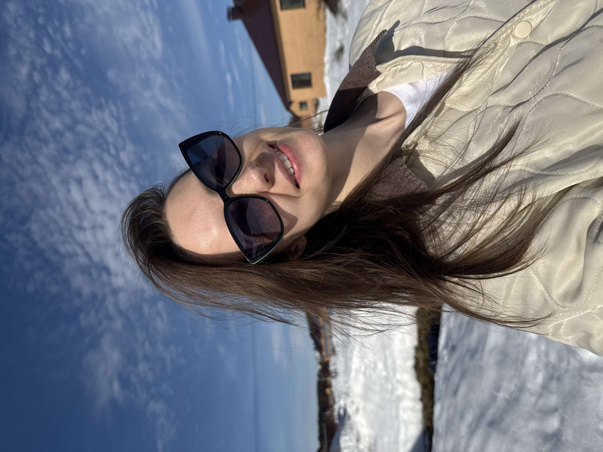 A female individual is depicted in a close-up, selfie-style photograph, outdoors in Chaikovsky, Russia. She has long, dark brown hair, which appears wind-blown, and is wearing black cat-eye sunglasses. Her facial expression is a smile, with upper teeth visible. She is dressed in a light-colored, possibly beige or cream, quilted jacket featuring a brown corduroy collar, worn over a white crew-neck t-shirt. The background is composed of a clear blue sky with scattered cirrus clouds. The ground is covered in snow, with patches of exposed brown earth visible. In the mid-ground, a detached house with yellow-orange siding and a reddish-brown sloped roof is present, with another similar structure partially visible further behind it. Utility lines are visible horizontally in the left portion of the upper frame. The scene is brightly illuminated by sunlight.