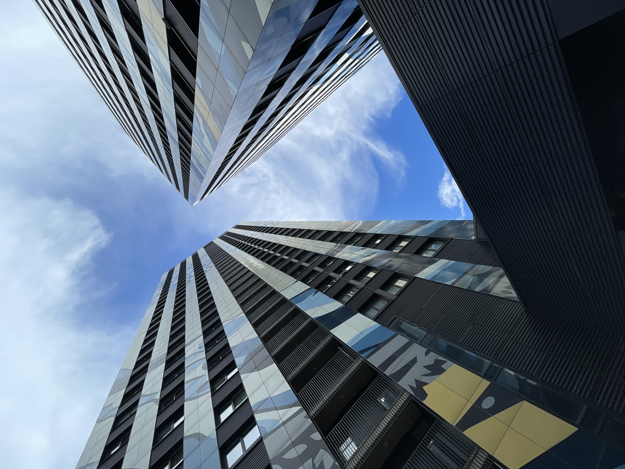 The image depicts a low-angle, upward view of modern architectural structures against a bright blue sky with scattered white clouds. Two tall, narrow buildings with distinctive patterned facades dominate the frame. The facade of the building on the left features horizontal bands of white and light gray panels interspersed with dark gray corrugated sections and sections with abstract blue and gray patterns. Some windows and balconies with metal railings are visible. The building on the right has a facade of reflective glass panels arranged in horizontal stripes, giving it a sleek, modern appearance. In the foreground, a dark, corrugated structure extends across the bottom of the image, partially obscuring the view of the buildings. The overall scene conveys a sense of urban scale and contemporary design. There are no people or animals visible in the image. The architectural style and material choices are characteristic of modern urban development. The specific location of Moscow, Russia, is not evident from the visual information provided in the image.