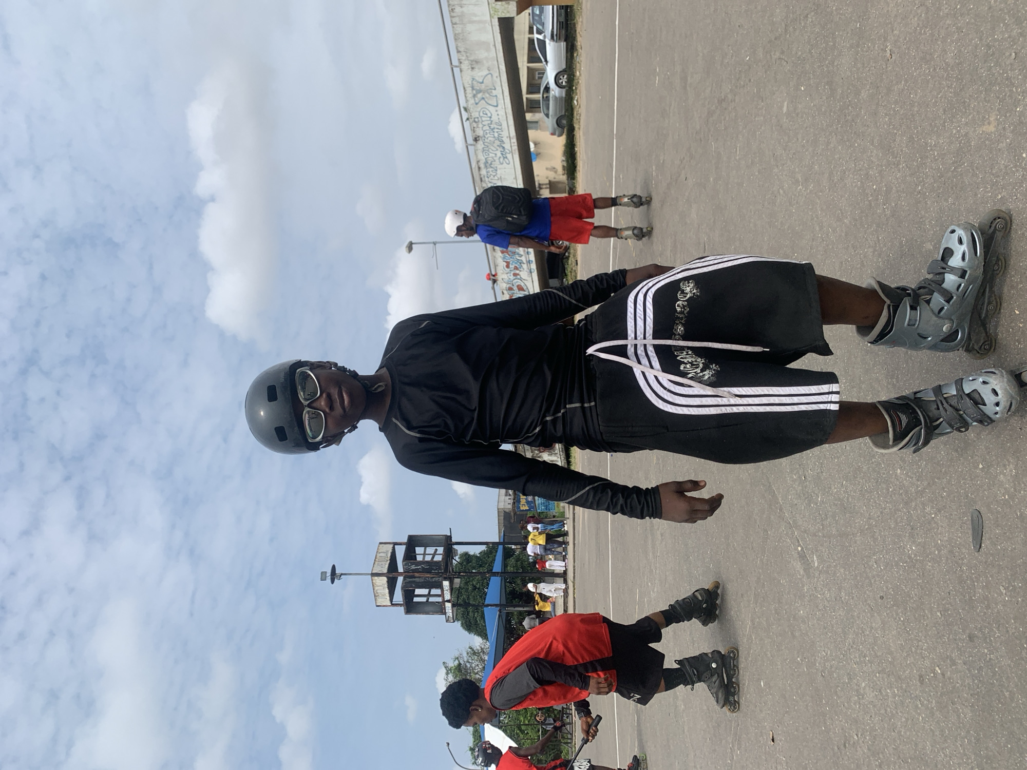 In Alagbede, Nigeria, on an outdoor paved surface, a young person wearing a black long-sleeved shirt, black shorts with white stripes, a black helmet, and rollerblades stands facing the camera. The person is wearing sunglasses. To the left, another young person in a red sleeveless shirt over a gray long-sleeved shirt and black shorts on rollerblades is also visible, holding a black stick. In the background, several other individuals are present. A concrete structure with platforms and a tall pole stands in the mid-ground, partially obscured by trees. Further in the background, a bridge with graffiti can be seen, and cars are parked. The sky is partly cloudy.