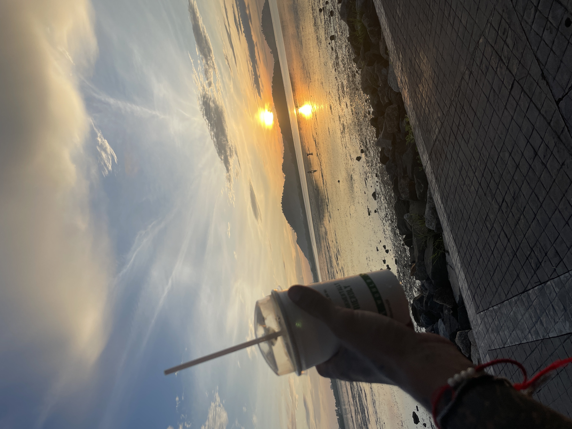 In Phuket City Municipality, Thailand, a hand holds a white disposable cup with a straw against a backdrop of a serene sunrise over a body of water. The sun, a bright orb, casts a warm golden glow across the sky and reflects on the water's surface, creating a shimmering path of light. Silhouetted against the horizon are distant hills. The foreground features a paved walkway and a rocky shoreline. The cup has text printed on it that appears to say "ENVIRONMENTALLY FRIENDLY". There are two small figures visible in the distance walking near the water's edge.