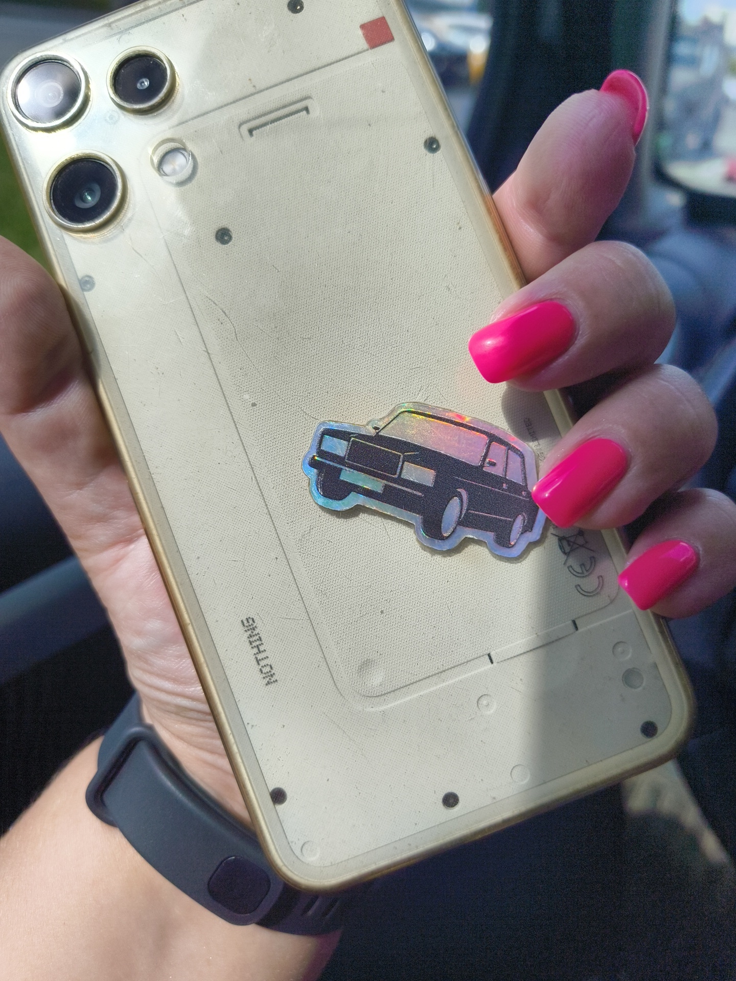 A close-up shot focuses on the back of a gold-colored smartphone, held by a person with bright pink fingernails. The phone has a textured back with visible screws and a small, holographic sticker of a car. The brand name "NOTHING" is subtly printed on the back. A dark grey fitness tracker is worn on the wrist holding the phone. The background is blurred, suggesting an indoor setting, possibly a car interior, with hints of daylight. The image does not contain any identifiable information about the city of Moscow, Russia.