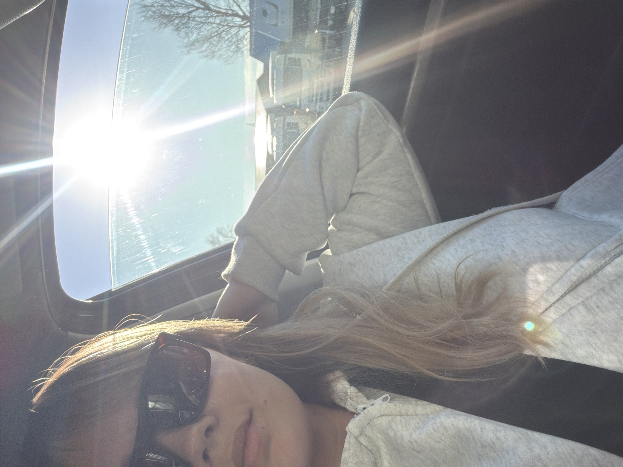 A young woman wearing sunglasses and a light gray hooded sweatshirt is in a car. The bright sun is shining through the sunroof. The location cannot be definitively determined as Chaikovsky, Russia without further context.