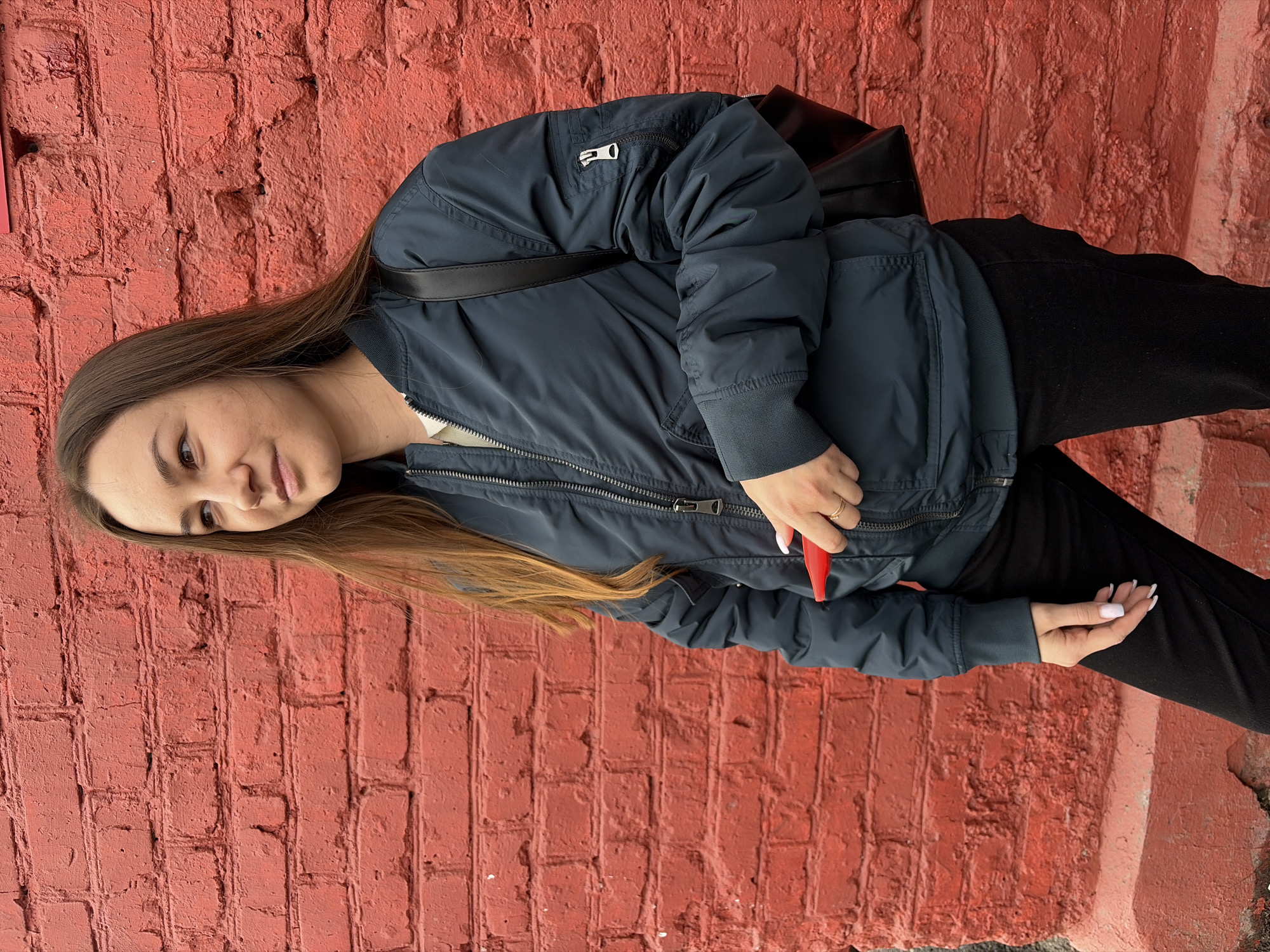A young woman with long brown hair stands against a red brick wall. She wears a dark blue bomber jacket over a white top and black pants. A black shoulder bag is slung over her shoulder. She is holding a small red object in her left hand. The background consists entirely of the red brick wall. The location is Izhevsk, Russia.