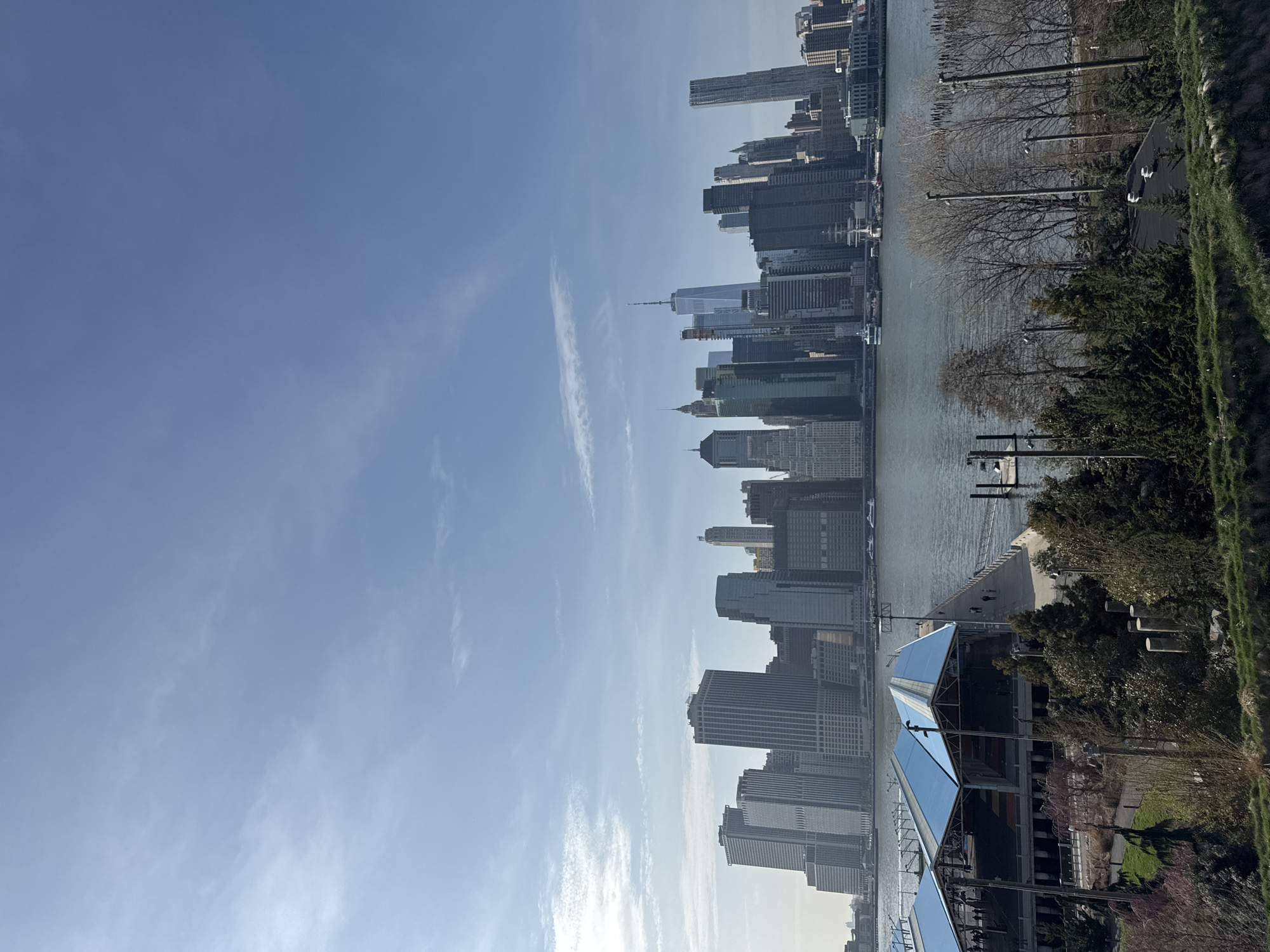 This image shows the New York City skyline from across the East River, likely from Brooklyn. The buildings include One World Trade Center. The photograph appears to have been taken on a sunny day with a clear, blue sky. The East River is visible with piers. Part of a building with a reflective roof is at the bottom of the frame. Bare trees and green grass are on the Brooklyn side of the river.