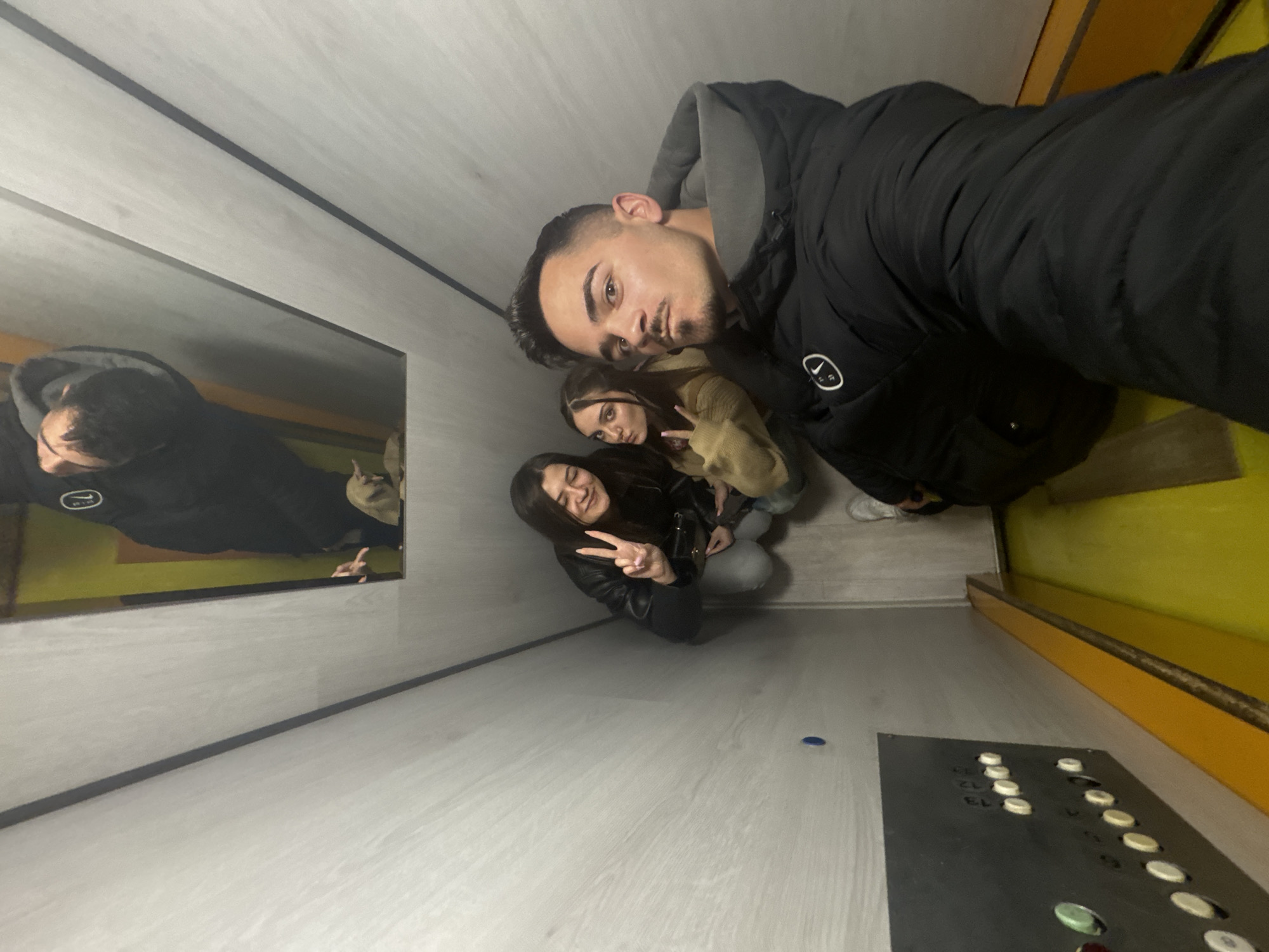 The photograph, taken from an elevated perspective, depicts three individuals inside an elevator. A man in a dark jacket with a gray hood is closest to the camera, taking the selfie. Two women are positioned behind him, both making peace signs with their hands. One woman is wearing a black leather jacket, and the other is wearing a tan-colored jacket.
The elevator's interior is constructed with light gray panels. To the left, a mirror reflects the scene. On the right, there is a yellow accent panel, and at the bottom, a dark panel with elevator floor buttons. The buttons have floor numbers near them. A small blue object is also visible on the floor.
The location cannot be determined to be in Sofia, Bulgaria, or any other specific city without further information.