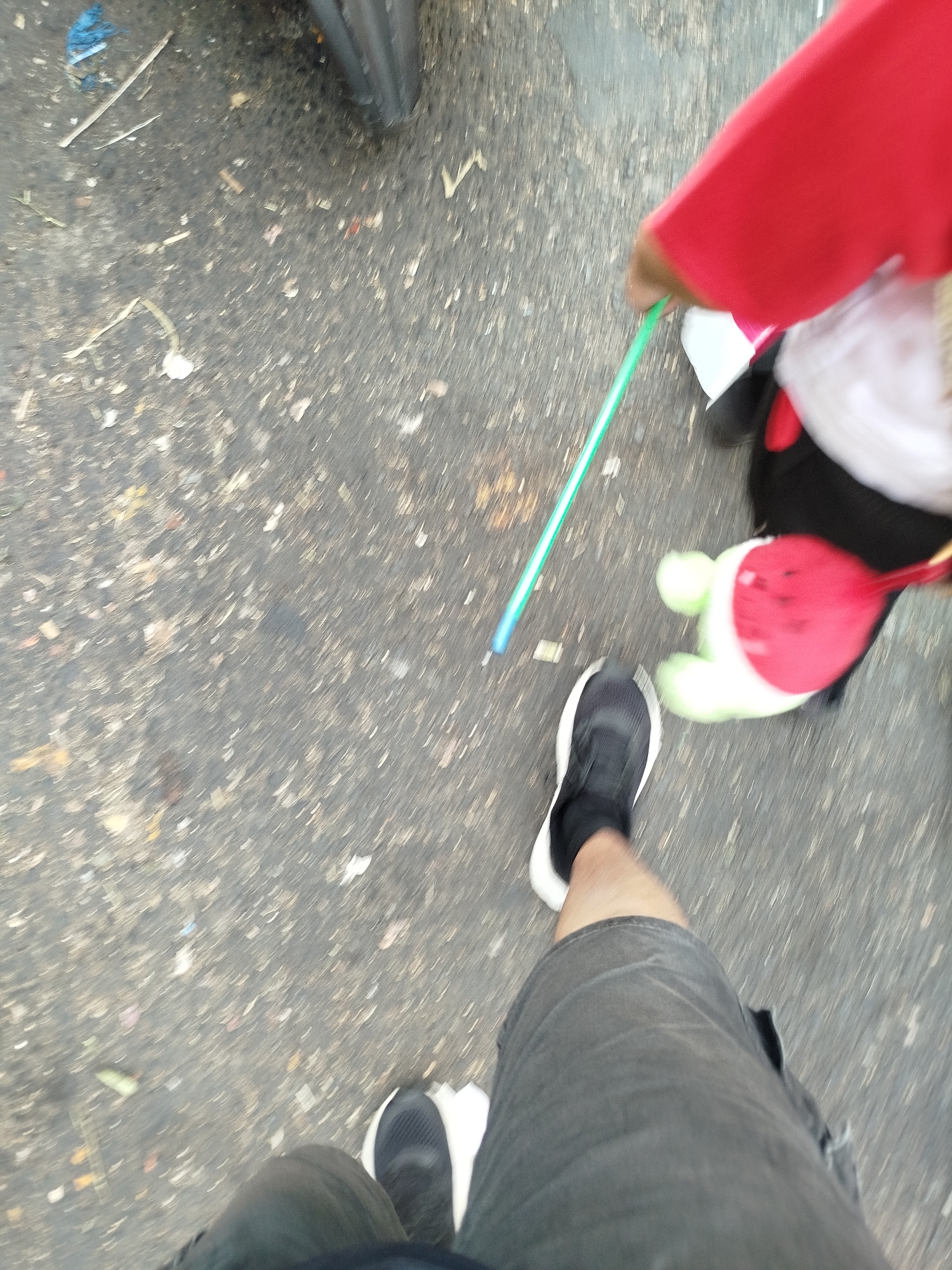 The image depicts a ground-level view of a person's legs and feet, wearing black athletic shoes with white soles and dark grey shorts. The person is standing on a textured asphalt surface littered with small debris. To the right, a child is visible, wearing a red garment and holding a thin, green, rod-like object. The child is also interacting with a stuffed toy that has light green limbs and a red and white body with red heart-like markings. The overall impression is of an outdoor scene, possibly a street or sidewalk, with a motion blur suggesting movement. The location is specified as the city of Surabaya, Indonesia, though the image itself does not contain any explicit visual cues to confirm this.
