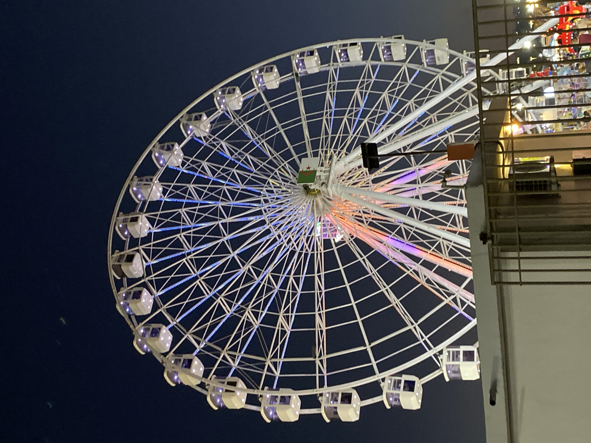 In the city of Hussein Dey, Algeria, a large Ferris wheel is illuminated against a dark night sky. The wheel is adorned with white gondolas and is outlined with blue and pink lights, creating a vibrant spectacle. The structure is supported by white spokes that radiate from the center, and at the hub of the wheel, the Algerian flag is prominently displayed. In the foreground, a white structure with a metal fence partially obscures the view of the Ferris wheel's base and the surrounding amusement park area, which appears to be bustling with activity and lit with various lights. The overall scene depicts a lively evening at an amusement park.