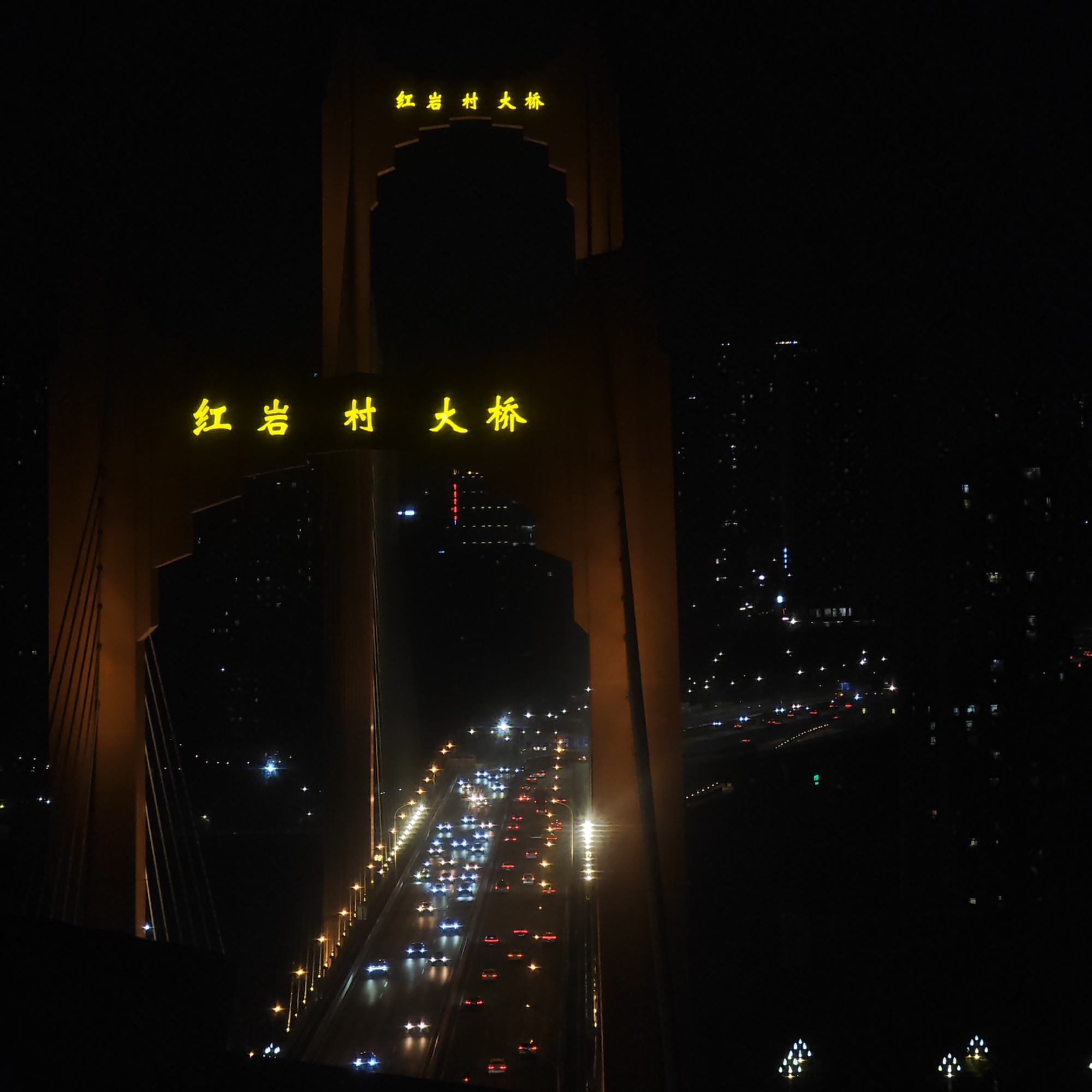 An eye-level night shot of Hongyancun Bridge in Yuzhong District, China, features illuminated yellow Chinese characters at the top and mid-section. A darkened bridge spans the lower part, with streams of car headlights and taillights running across. Tall buildings on either side fade into the darkness.