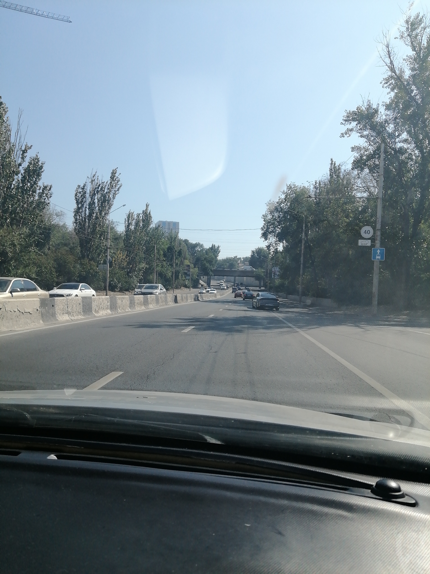 The image depicts a view from inside a vehicle traveling on a road in Rostov-on-Don, Russia. The road is a multi-lane highway with concrete barriers separating the traffic lanes from the adjacent embankment. Trees line the embankment, and a traffic sign indicating a speed limit of 40 km/h is visible on the right side of the road. Several vehicles are driving on the highway in the same direction as the camera. The sky is clear and blue, suggesting fair weather. The foreground shows the dashboard and windshield of the vehicle from which the photo was taken.