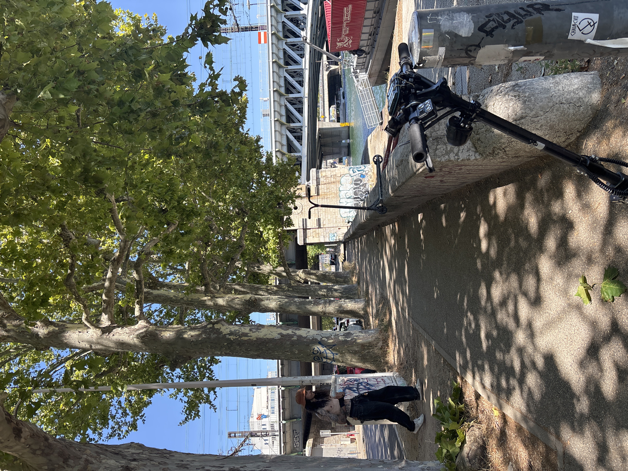 An electric scooter is parked on a concrete pathway next to a low stone wall. The pathway is lined with large trees, casting dappled shadows on the ground. A person wearing a brown hat and dark pants stands next to a utility box, looking towards the right of the frame. Beyond the pathway, a river is visible, with bridges and train tracks spanning across it. In the background, a red shipping container is docked. The scene appears to be an urban area in Lyon, France, possibly along the Rhône River.