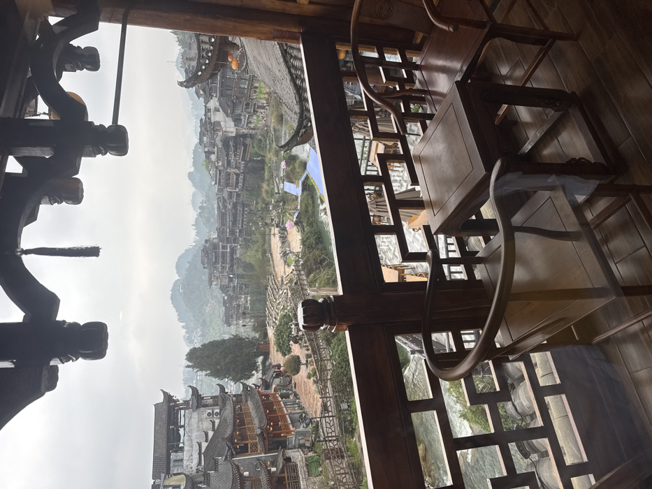 The image displays an elevated view of Furong Ancient Town, China, under an overcast sky. In the foreground, a dark wooden balcony features a geometrically patterned railing, two traditional dark wooden chairs with rounded backs, and a square table, all situated on a tiled floor. Above, an ornate carved wooden ceiling structure with a dangling black tassel frames the top of the scene.
Below the balcony, a river with visible rapids flows through the town. Its banks are lined with traditional Chinese buildings, characterized by dark tiled roofs, multi-tiered wooden structures, and open balconies. Several red lanterns are suspended from these buildings. On the left bank of the river, a stone amphitheater with tiered seating is visible, alongside a prominent tall conifer tree. Winding pedestrian paths and bridges connect various sections of the town. On the right side of the river, a bright blue pathway or structure is situated near large pink floral sculptures. Several indistinct human figures are observed on the pathways and in the open areas.
The town's architecture extends up the surrounding forested hillsides, with more traditional buildings nestled among the trees. Distant mountains are visible through a pervasive atmospheric haze, indicating an overcast day.