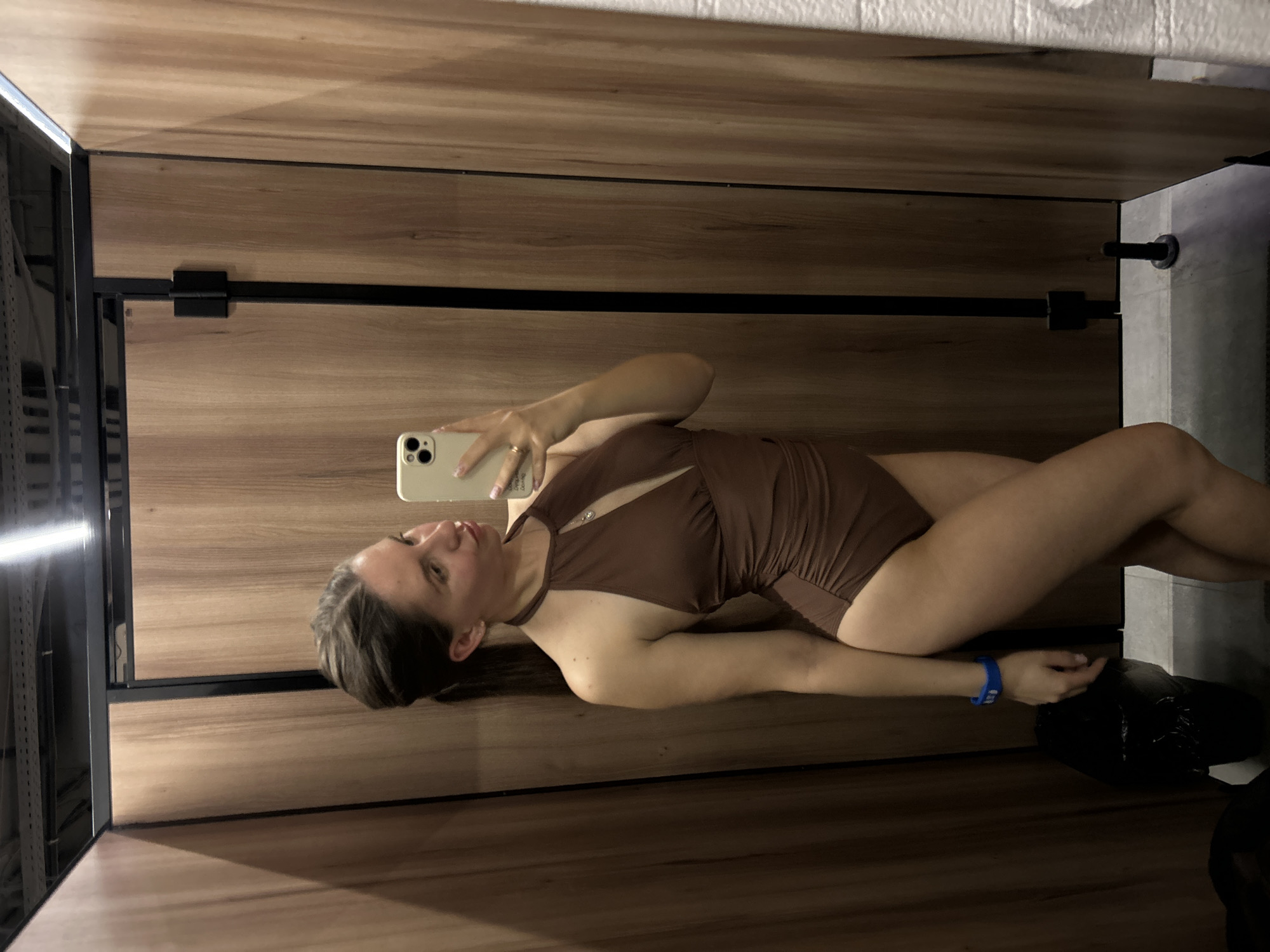 The image displays a young woman taking a mirror selfie in what appears to be a changing room or locker room. She is positioned as if lying down or leaning back against a surface, capturing her reflection and the surrounding environment. The woman wears a brown, one-piece swimsuit or leotard featuring subtle ruched details on the bodice. Her dark hair is pulled back, and she holds a light-colored smartphone, possibly an iPhone, in her right hand, which also sports a ring. A blue wristband with indiscernible white text or numbers is visible on her left wrist, suggesting she is at a public facility like a gym, spa, or swimming pool.
The background is characterized by wooden paneled walls with prominent dark, rectangular metal framing or shelving units. In the far left of the image, part of a metallic shelving system or locker array can be seen. A white, textured surface, possibly a ceiling or an adjacent wall, is visible in the upper right. A dark bag or object rests on the surface near her feet on the right side. The overall lighting is artificial and indoor. The setting is in Tver, Russia.