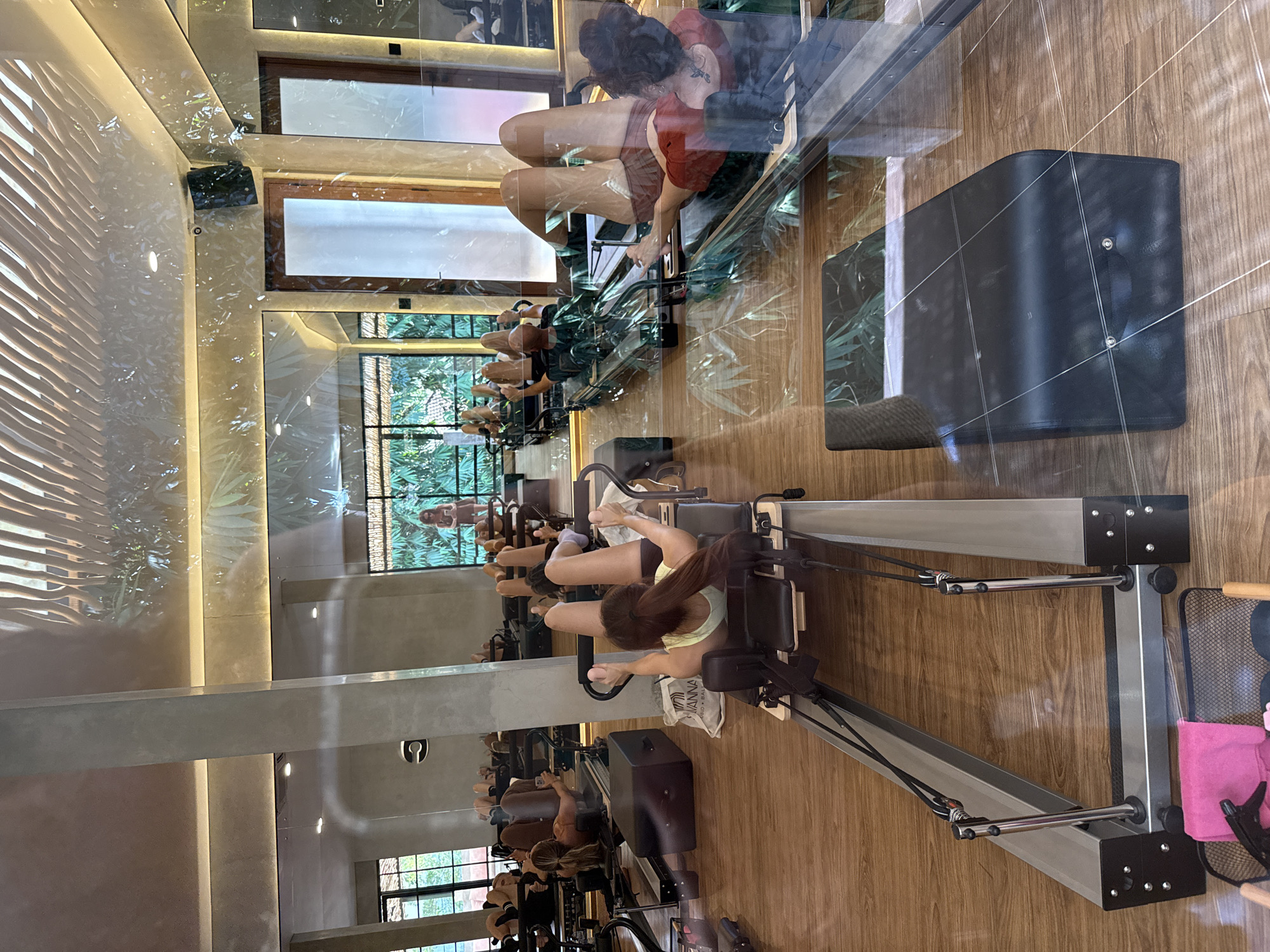 In a dimly lit studio with blonde wood floors, several women are performing Pilates on reformers, which are exercise machines designed for resistance training. The studio features large windows looking out onto lush greenery, suggesting a location in a tropical or subtropical area. The architecture includes exposed concrete pillars and a ceiling with an artistic, wave-like design. The overall ambiance is one of a high-end fitness or wellness center. The scene is captured through a reflective surface, likely glass, which distorts and overlays reflections of plants and the ceiling design onto the foreground. The specific location in the city of Ubud District, Indonesia, is suggested by the lush vegetation visible through the windows.