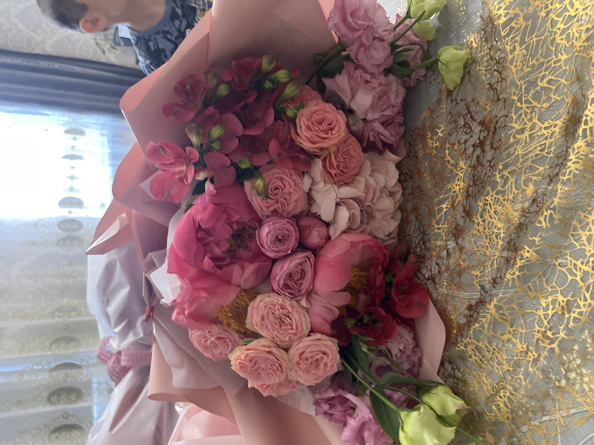A bouquet of pink and red flowers is featured prominently. The bouquet is wrapped in pink paper, and includes roses, peonies, alstroemeria and other blooms. Parts of a child can be seen in the top left corner. The background includes a gold and silver patterned wall. The location is Kaspiysk, Russia.
