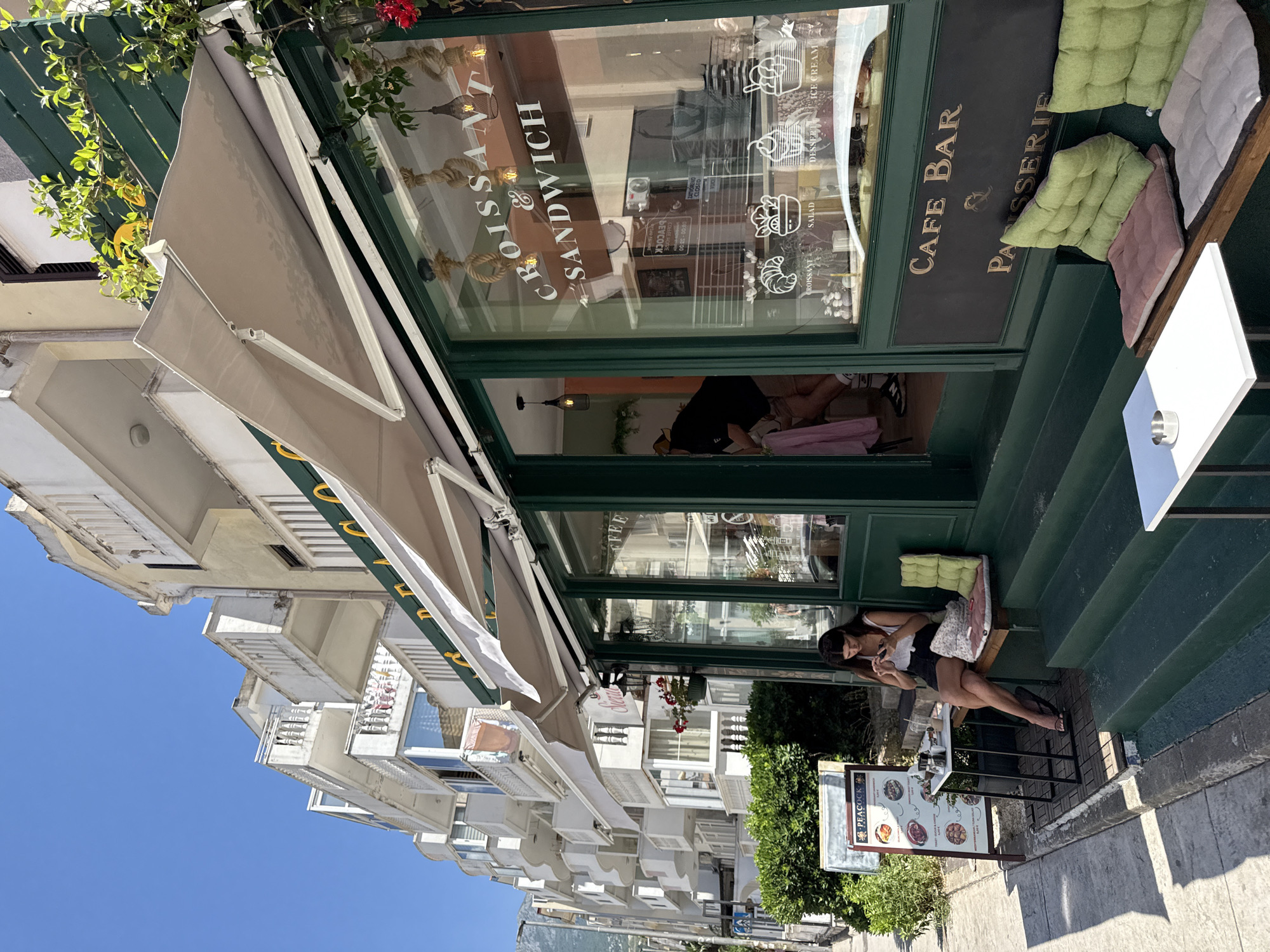 In Budva, Montenegro, a woman is seated on a bench outside a cafe with green trim and large windows. She is looking down at her phone and has a drink on a small table next to her. The cafe has awnings extended over the windows, and a sign in the window reads "CROISSANT & SANDWICH" and "CAFE BAR & PATISSERIE." Another sign displays food items with prices. The street is lined with buildings with white balconies, and lush green bushes are visible. The sky is clear and blue.