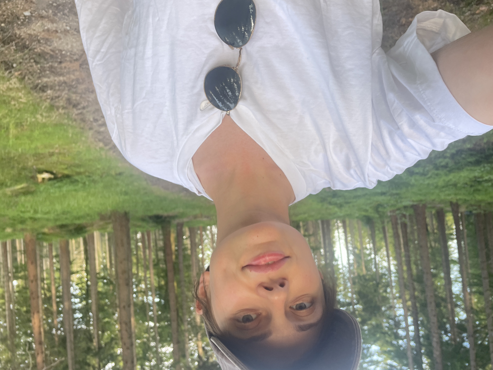 A young woman with short brown hair, wearing a gray baseball cap and a white t-shirt, poses for a selfie in a forest. Sunglasses are hanging from the neckline of her shirt. She is standing on a dirt path with green grass and tall, slender trees surrounding her. The scene is bathed in natural light, suggesting it is daytime. The location is the city of Szklarska Poręba, Poland.