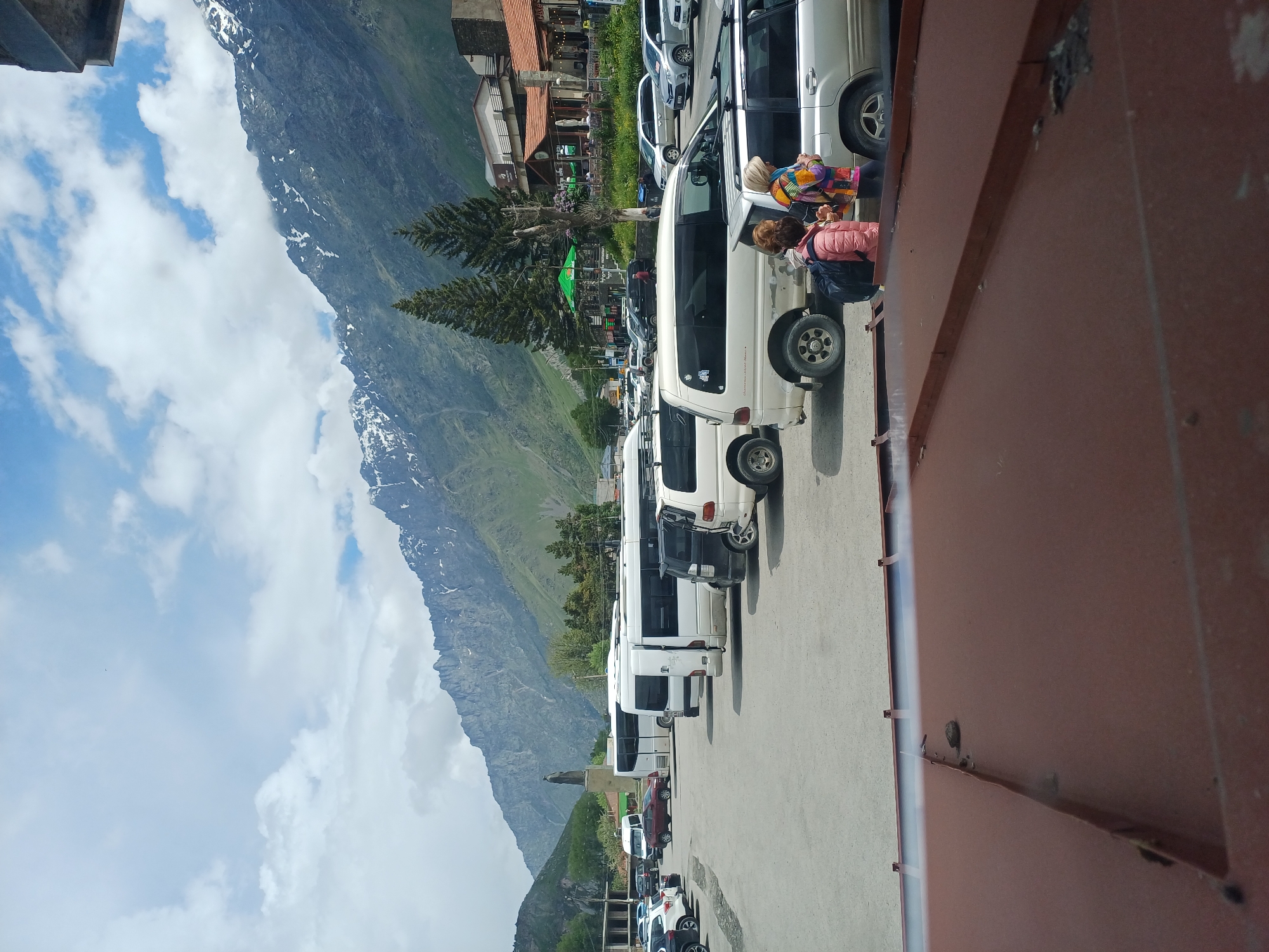 A wide-angle shot captures a parking lot filled with vehicles, including several white minivans and buses, as well as cars. In the background, a rugged mountain range with snow-capped peaks dominates the skyline under a partly cloudy sky. Two evergreen trees stand in front of a few buildings that appear to be part of a small town or tourist area, with a statue visible on a slight rise to the left. Two women are standing near a silver SUV in the foreground, one wearing a pink jacket and a backpack, the other in a colorful jacket. The image appears to have been taken from a vantage point slightly above the parking lot, possibly from a balcony or elevated structure, as a dark brown, corrugated metal surface is prominent in the lower portion of the frame.