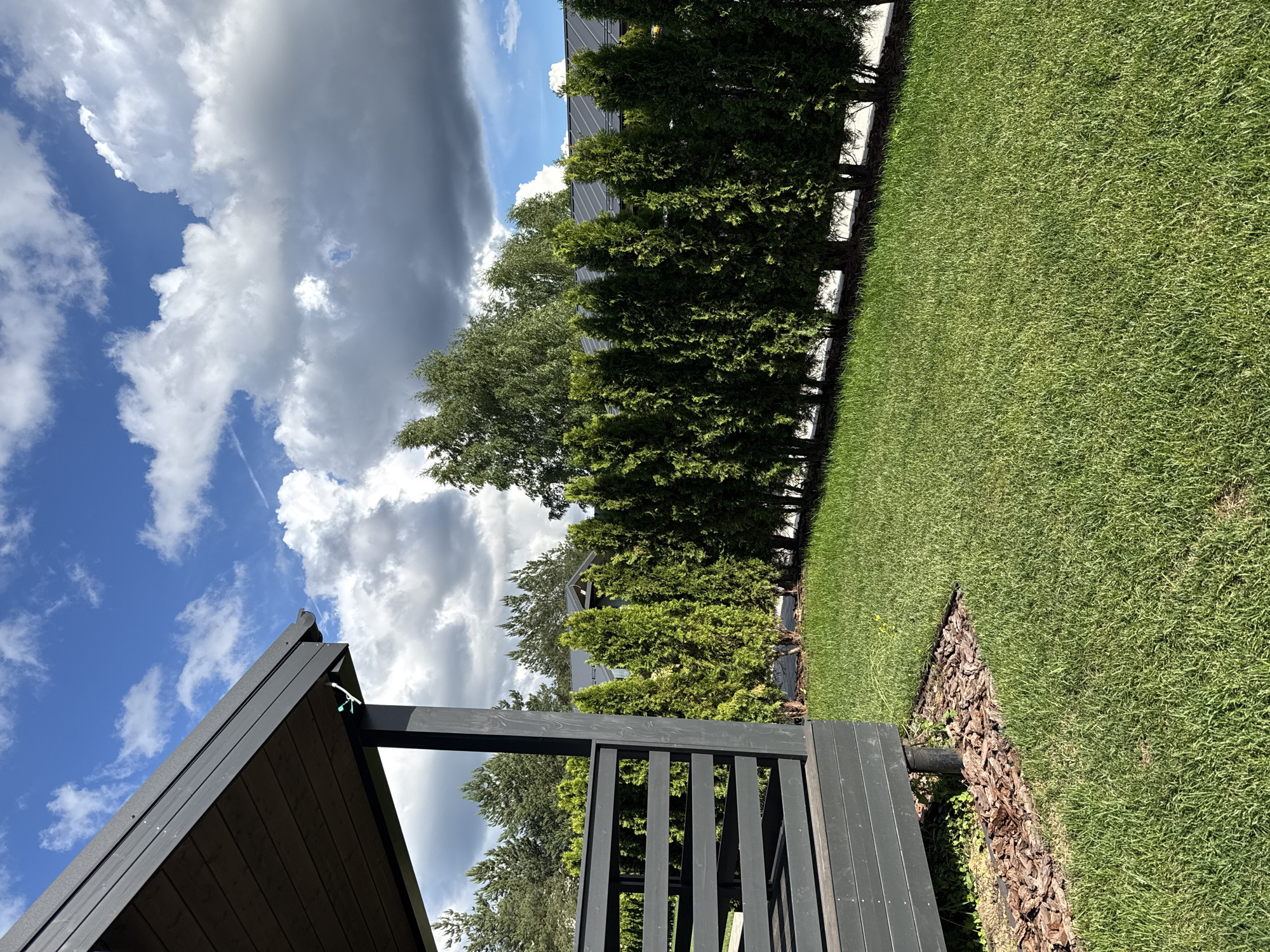 This is a high-angle, landscape orientation, outdoor photograph taken in Жостово, Russia. The scene depicts a backyard on a bright, partly cloudy day. The foreground features the dark wooden roof and railing of an outdoor structure, possibly a gazebo or pergola. Beyond this structure is a vibrant green lawn that extends to the right of the frame. A border of mulch and some low-lying greenery separates the lawn from a dense line of dark green evergreen trees. Behind the evergreens, a building with a dark roof is partially visible, suggesting a residential area. The sky is a prominent feature, with a mix of bright blue sky and dramatic, voluminous white and grey clouds. A thin contrail is visible in the upper left portion of the sky. The lighting suggests it is daytime, likely late morning or early afternoon, with the sun casting shadows. The overall mood is serene and highlights a well-maintained outdoor space. No people or text are visible in the image.