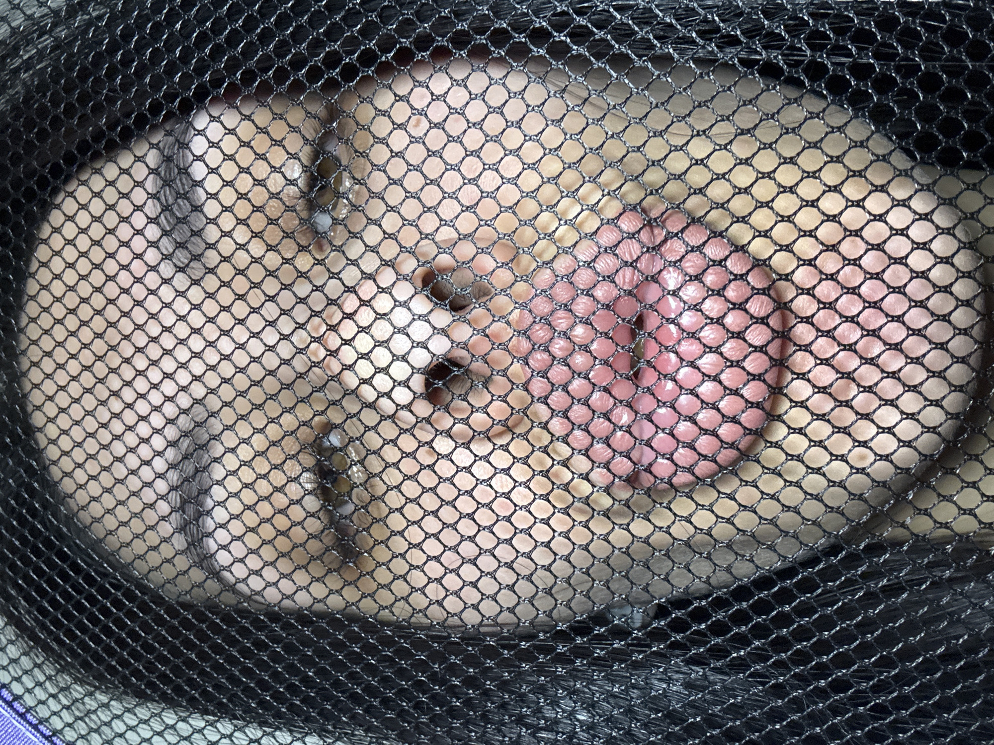 A close-up shot shows a human face obscured by a black mesh netting. The facial features, including the nose, mouth, and eyes, are visible through the netting. The skin tone appears light, with areas of redness around the mouth. The subject appears to be inside a mesh bag. The environment is not clearly discernible; however, based on the prompt, this could be in Votkinsk, Russia.