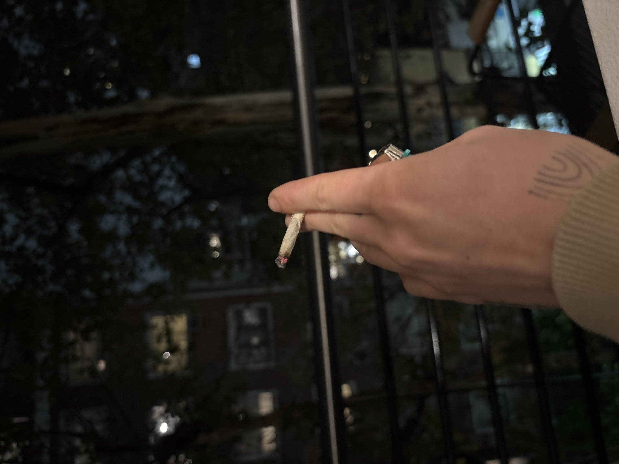 A hand holds a lit cigarette or joint outdoors at night. The hand has a tattoo on the wrist. The background features a window frame and a view of a building with lit windows, possibly in Buenos Aires, Argentina. The exact location in Buenos Aires is impossible to determine from this image alone.