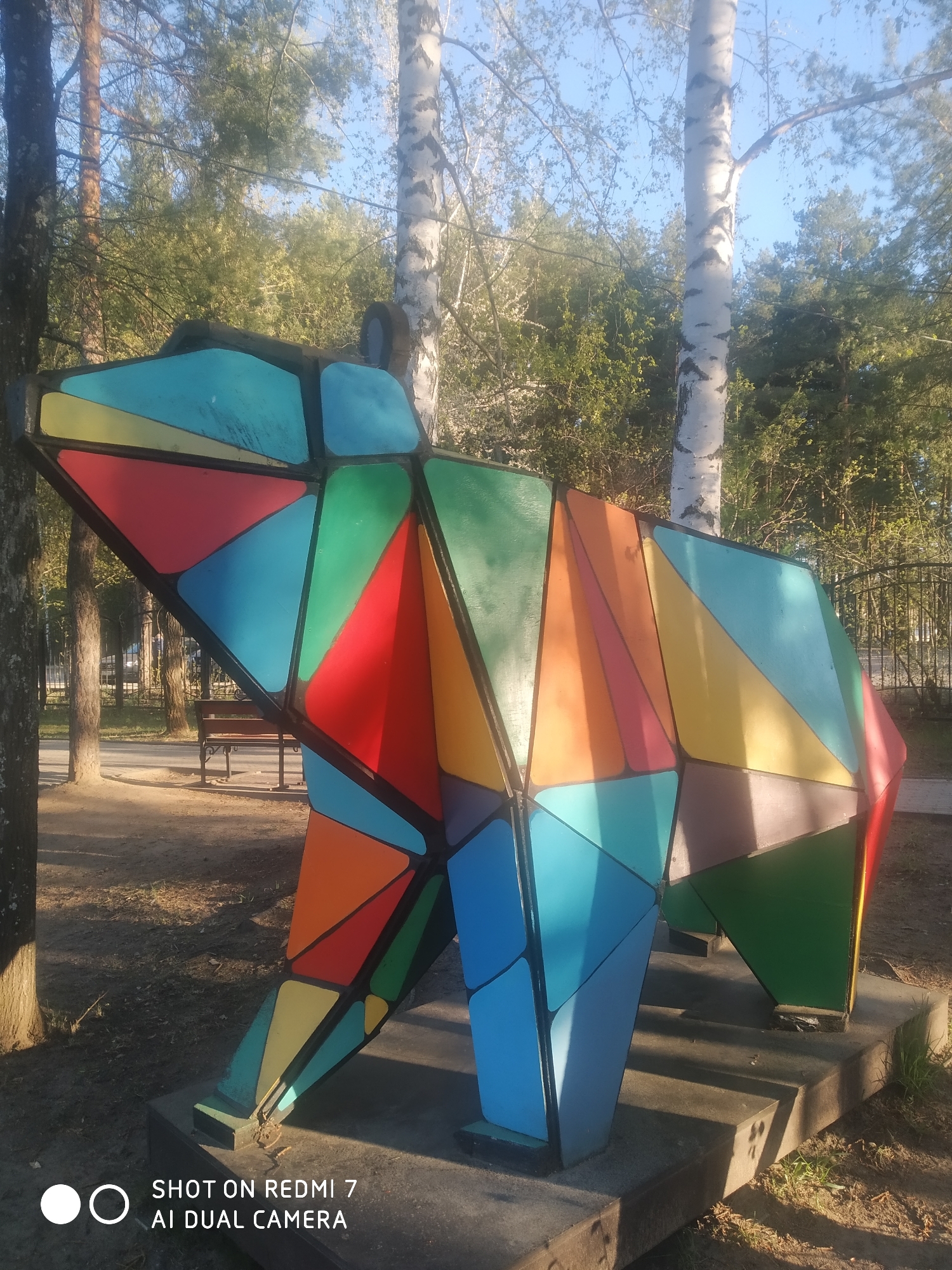 A brightly colored, geometric bear sculpture stands outdoors in Chaikovsky, Russia. The sculpture is constructed of various painted triangles in red, blue, green, yellow, and orange, creating a patchwork effect. It's positioned on a concrete base. Behind the sculpture, the scene features trees, including birch trees with white bark, and a bench is visible in the background. The photo was taken on a sunny day.