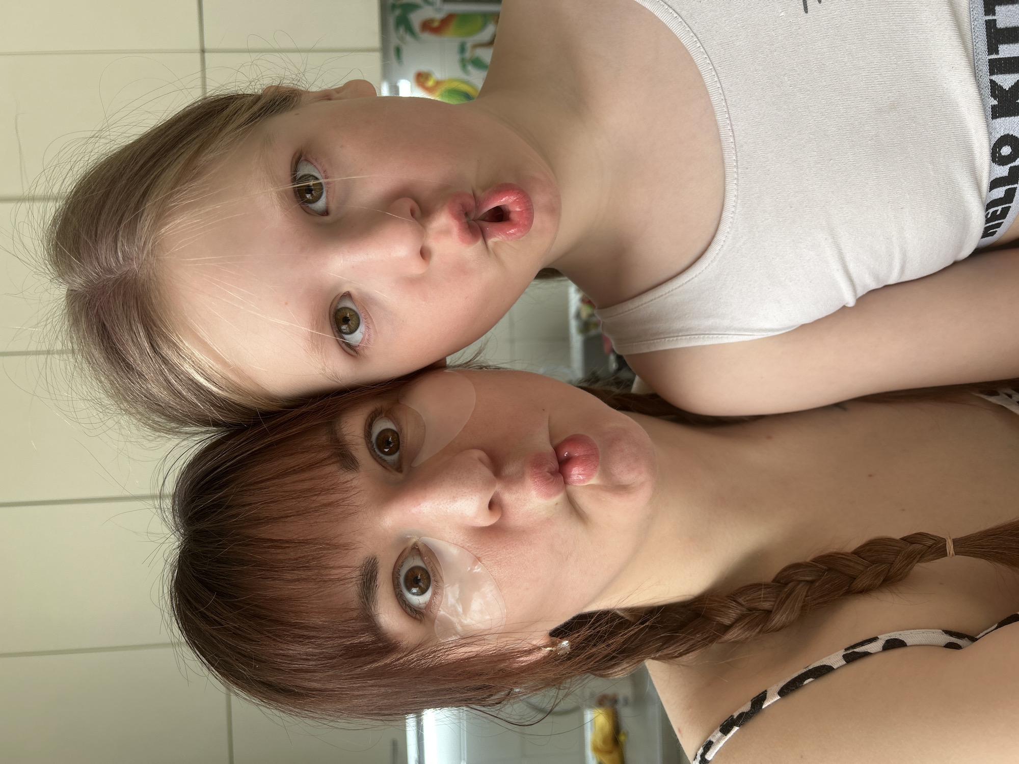 This image depicts two females, an adult and a child, captured in a close-up, chest-up shot within a domestic interior in the city of Фоки, Russia. Both individuals are looking directly at the camera with wide eyes and their lips pursed into an exaggerated "fish face" expression.
The adult female, positioned on the left, has dark brown hair with bangs across her forehead and a visible braid extending over her left shoulder. She has two translucent, gel-like patches applied under her eyes. A small, clear stud earring is visible in her left ear. She is wearing a black and white animal print strap top.
The child female, positioned on the right, has light-colored hair. She is wearing a light cream or beige ribbed tank top. A grey elastic band with "HELLO KITTY" text in black is partially visible below the hem of her top.
The background consists of light-colored walls or cabinetry. On the right side, a decorative element featuring colorful parrot or tropical bird imagery is partially visible. The lighting is bright and even.