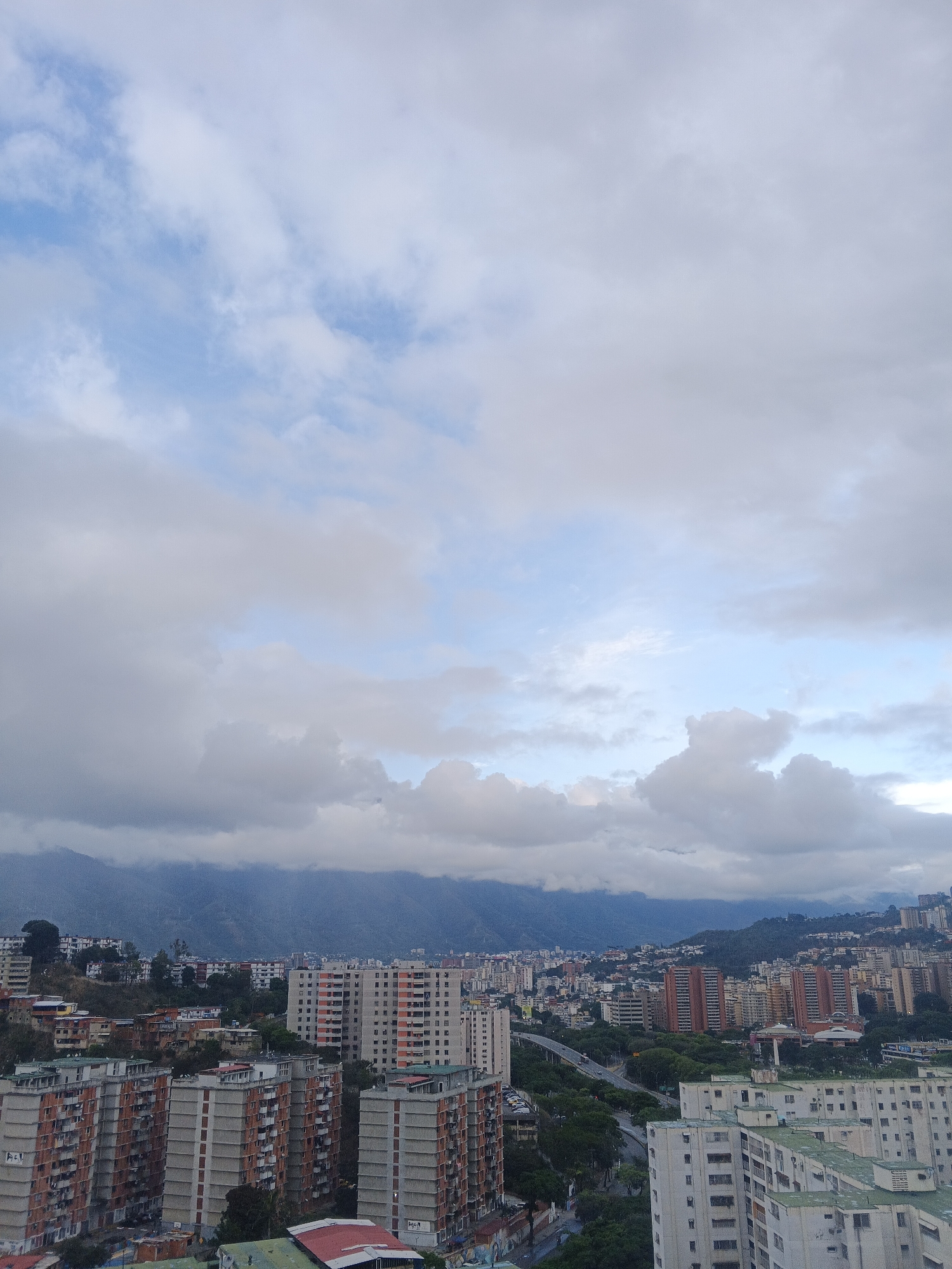 The image shows a cityscape of Caracas, Venezuela, under a sky filled with clouds. Mountains are visible in the background, partially obscured by the clouds. In the foreground, there are numerous high-rise buildings, primarily residential, with varying colors and heights. A major highway runs through the city, lined with trees and some vehicles. The overall scene appears overcast.