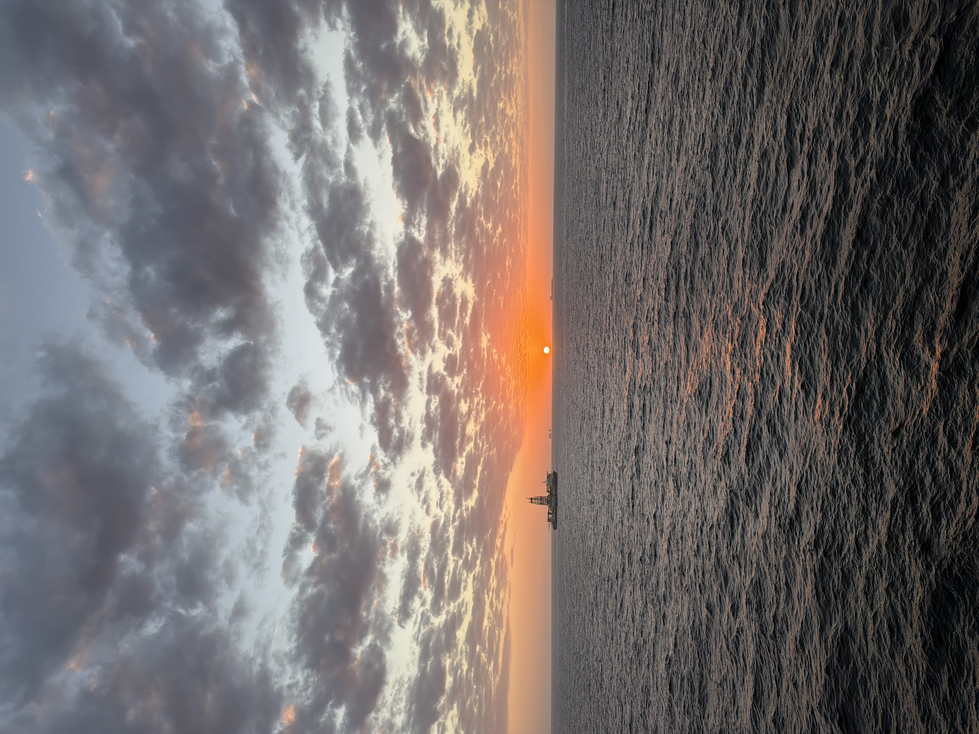 The image depicts a sunset over the ocean. The horizon is bisected, with a cloudy sky on the left and the ocean on the right. The sun is visible on the horizon, and there appears to be an oil rig silhouetted against the sunset. Location: Unknown, Unknown.