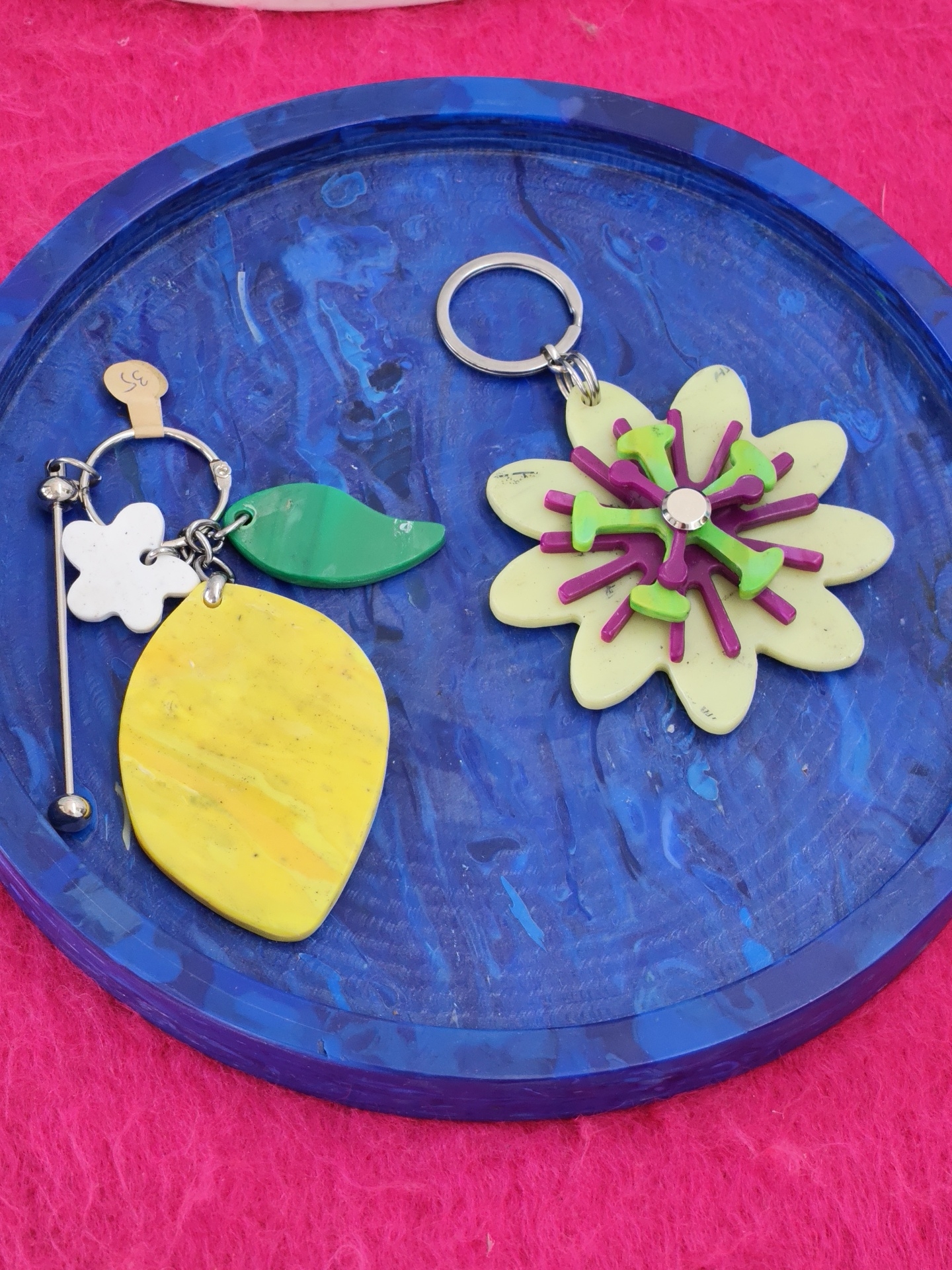 A close-up, top-down view shows two colorful, handmade keychains resting on a swirling blue and black marbled tray, which sits on a vibrant hot pink textured surface. The keychain on the left features a silver ring with a tan paper tag displaying the number "35". Dangling from the ring are a white four-petaled flower charm, a bright green leaf-shaped charm, and a large, elongated yellow charm with streaks of orange and white, all connected by silver jump rings and a vertical silver bar with two small silver spheres at the bottom. The keychain on the right has a silver ring holding a layered, light yellow flower charm. This flower charm has a purple center and radiating purple and green accents forming a spiky, layered blossom. The keychains and tray are placed on a surface that appears to be a rough, fuzzy fabric. The scene is brightly lit, highlighting the textures and colors of the objects. The background is out of focus, suggesting the image was taken in a market or retail setting, possibly in Batumi, Georgia, where such artisanal crafts are commonly displayed for sale.